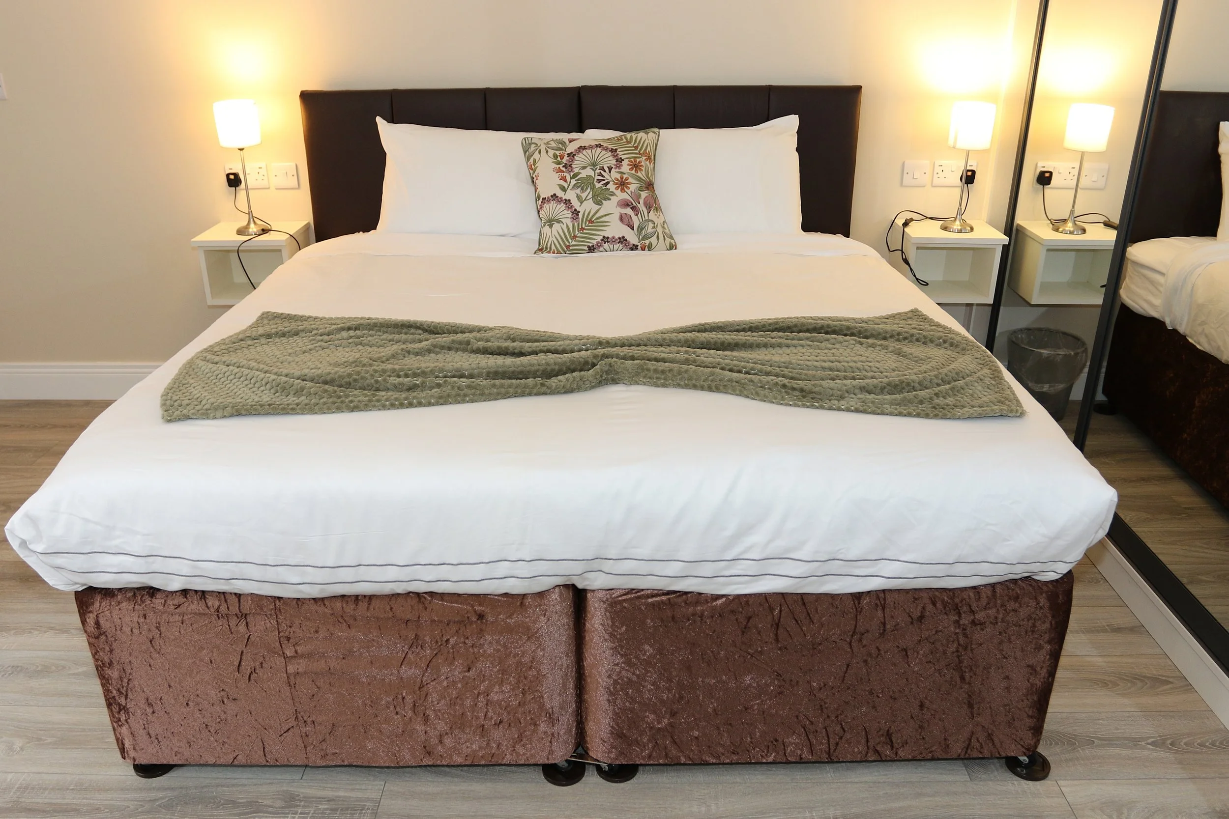A bedsheet with a brown base and a maroon bedframe, a bed with white sheets, a blanket, and a pillow with a floral pattern, located in a bedroom with two nightstands and lamps.