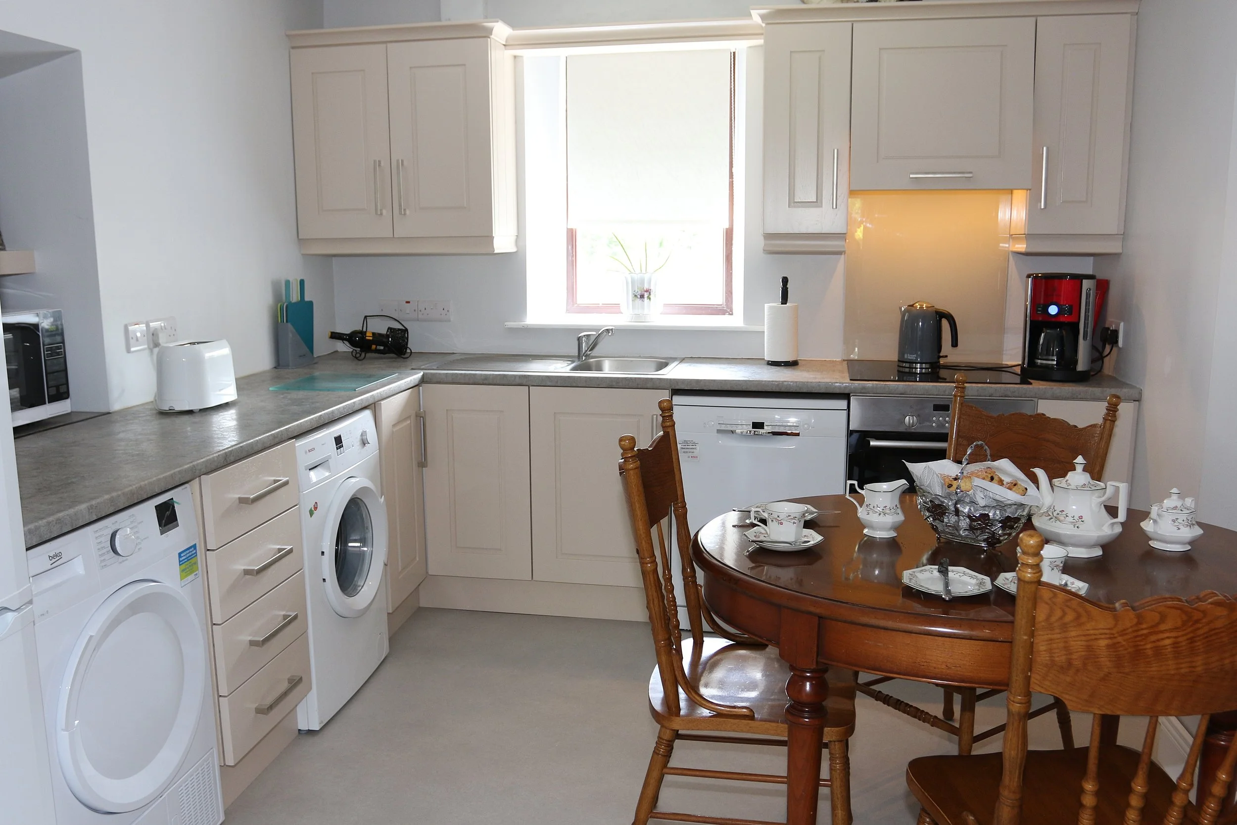 A kitchen with white cabinets, a window with a flowerpot on the windowsill, a countertop with various small appliances, a wooden dining table with chairs, and dishes set for a meal.