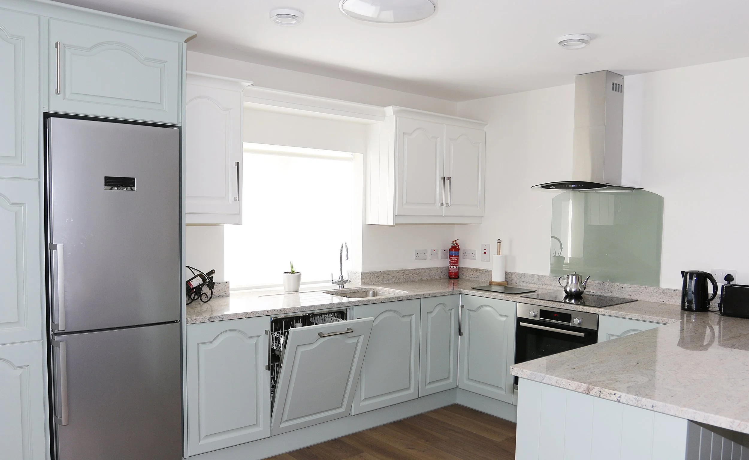 White kitchen with granite countertops, white cabinets, stainless steel refrigerator, black stove and oven, and kitchen appliances on the counter, including a kettle, toaster, and a dark-colored water bottle.