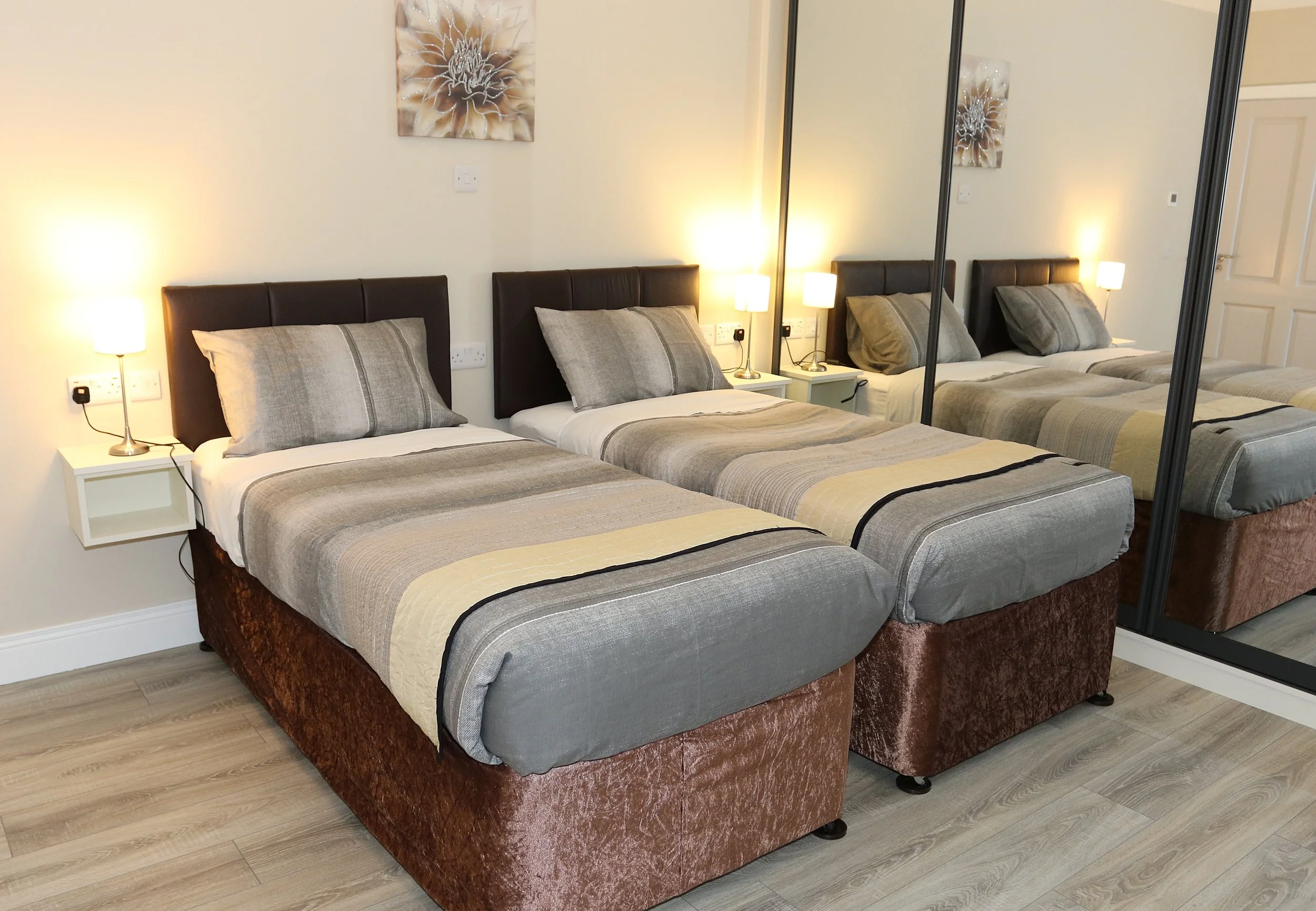 Three beds with brown headboards, beige and gray striped bedding, and pillows, flanked by identical white nightstands with lamps, a wall sculpture of a flower, and mirrored closet doors reflecting the room.