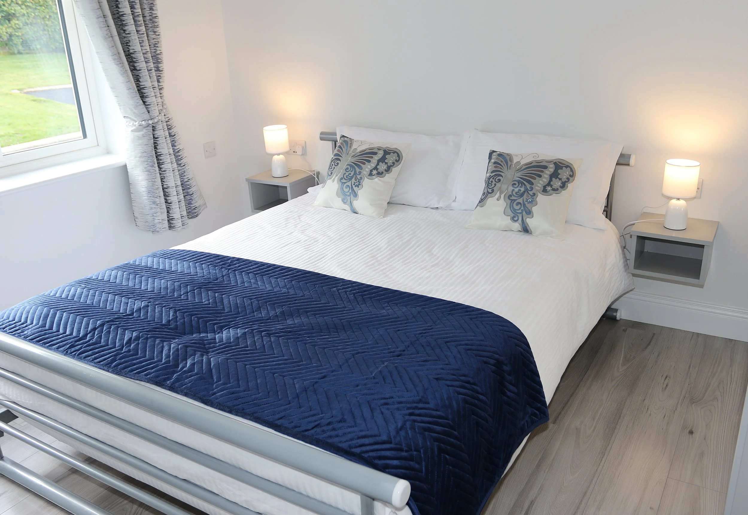 A neatly made bed with white bedding, blue quilt, and decorative pillows with butterfly patterns in a bedroom. Two white nightstands with lamps are on each side of the bed. A window with curtains showing a green outdoor scenery is on the left.