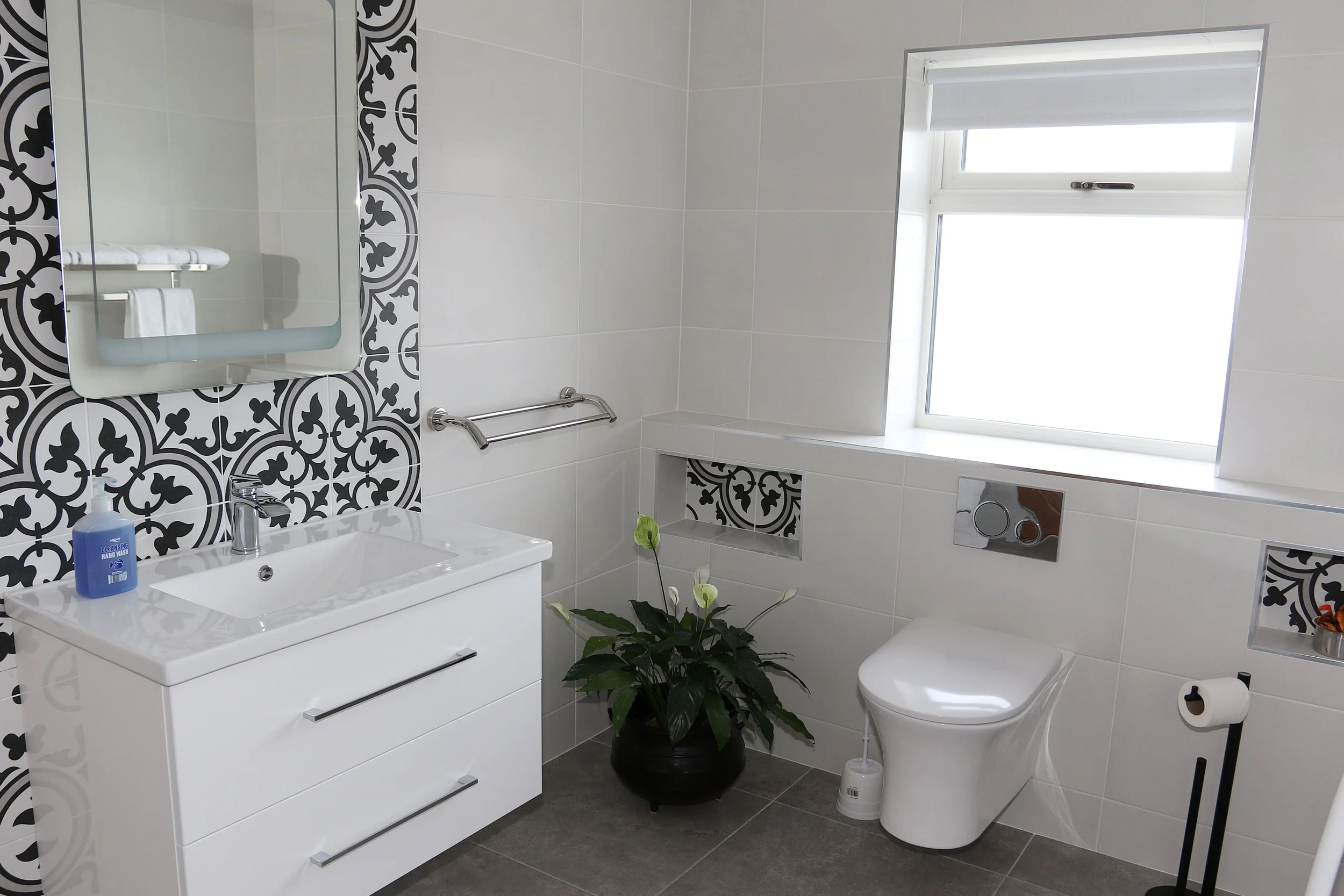A modern bathroom with white tiles featuring black and white patterned accents, a white vanity with a mirror, a toilet, a window with a white roller shade, green plants, a towel rack, and a soap dispenser.