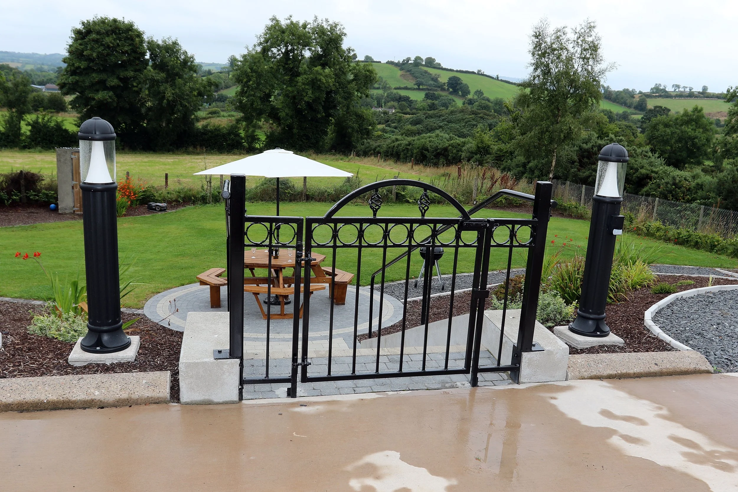 A backyard patio with a black wrought iron gate, a wooden picnic table with a large white umbrella, a paved circular area, and lush green landscape with rolling hills and trees in the background.