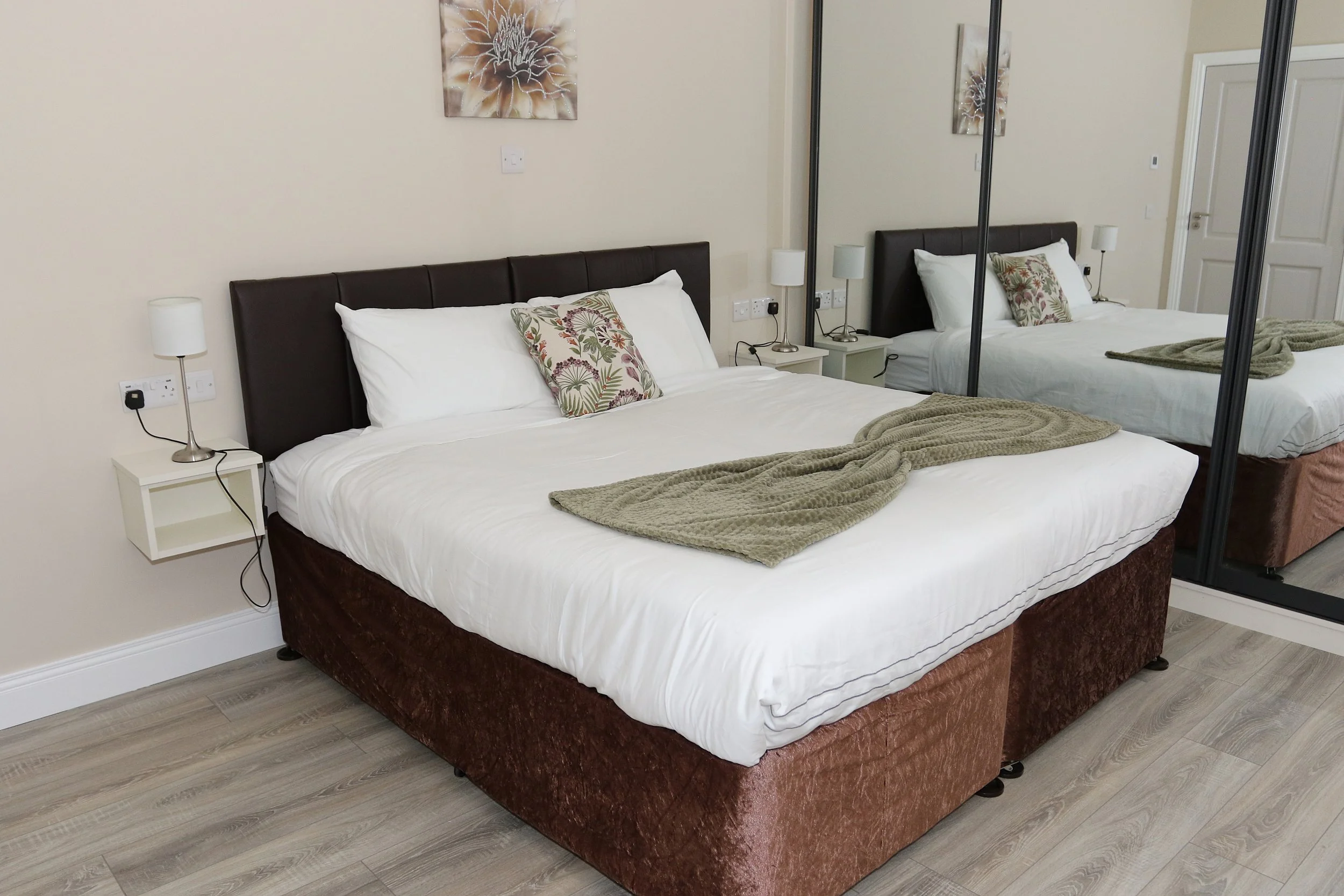 A bedroom with a double bed, two bedside tables with lamps, a mirror with sliding doors, wall art, and a light-colored wooden floor.