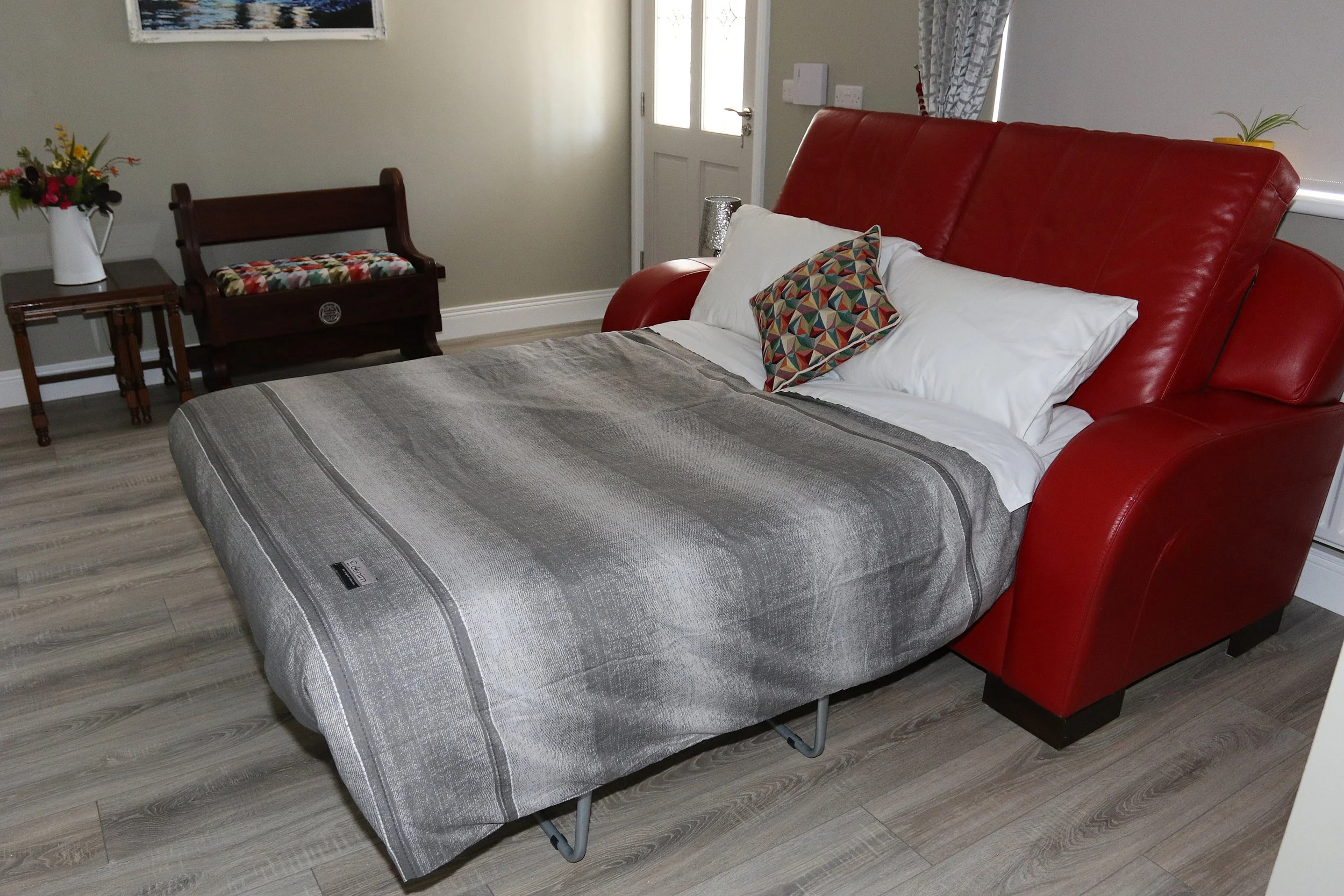 A bedroom with a double bed featuring a red headboard, white and patterned pillows, and a gray striped mattress. Next to the bed is a wooden bedside table with a white vase of colorful flowers. There is a wooden bench with a floral cushion near the door, which has a window. The floor is wood, and the wall is plain with a picture hanging.
