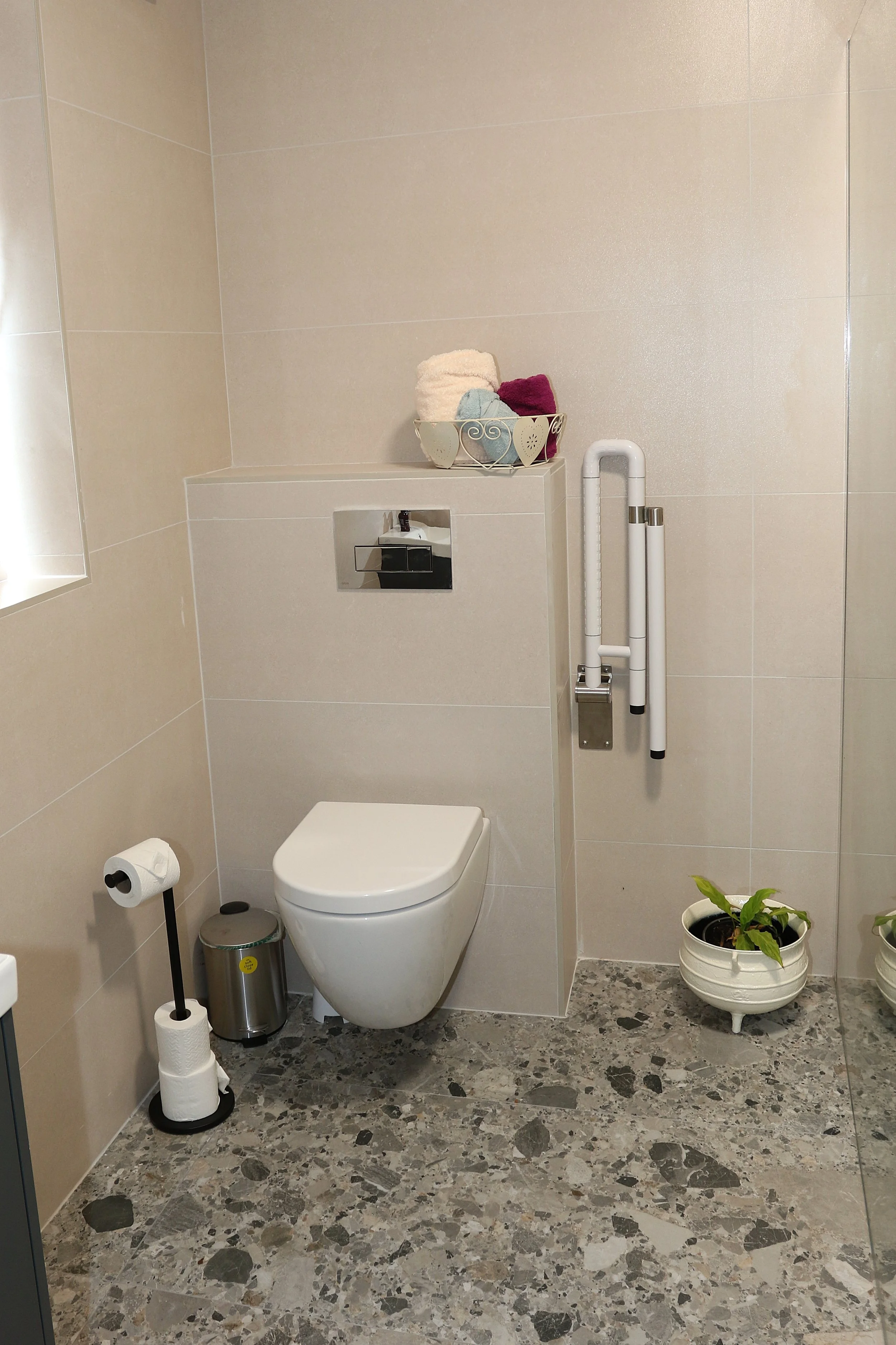 A modern bathroom with beige tiled walls, a white toilet, a toilet paper holder on a stand, a small trash can, and a potted plant on the floor. On the wall above the toilet, there is a shelf with rolled towels and decorative hearts.