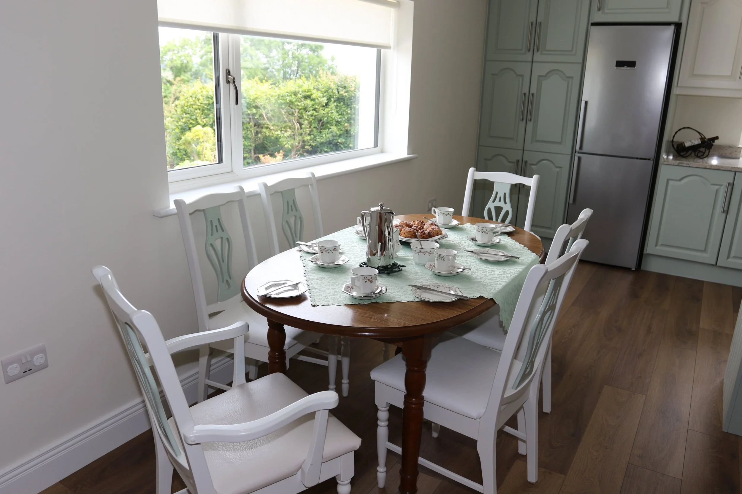 Dining area with a round wooden table set for tea with white china, a silver teapot, and pastries, surrounded by white chairs, in a kitchen with large window and light-colored cabinets.