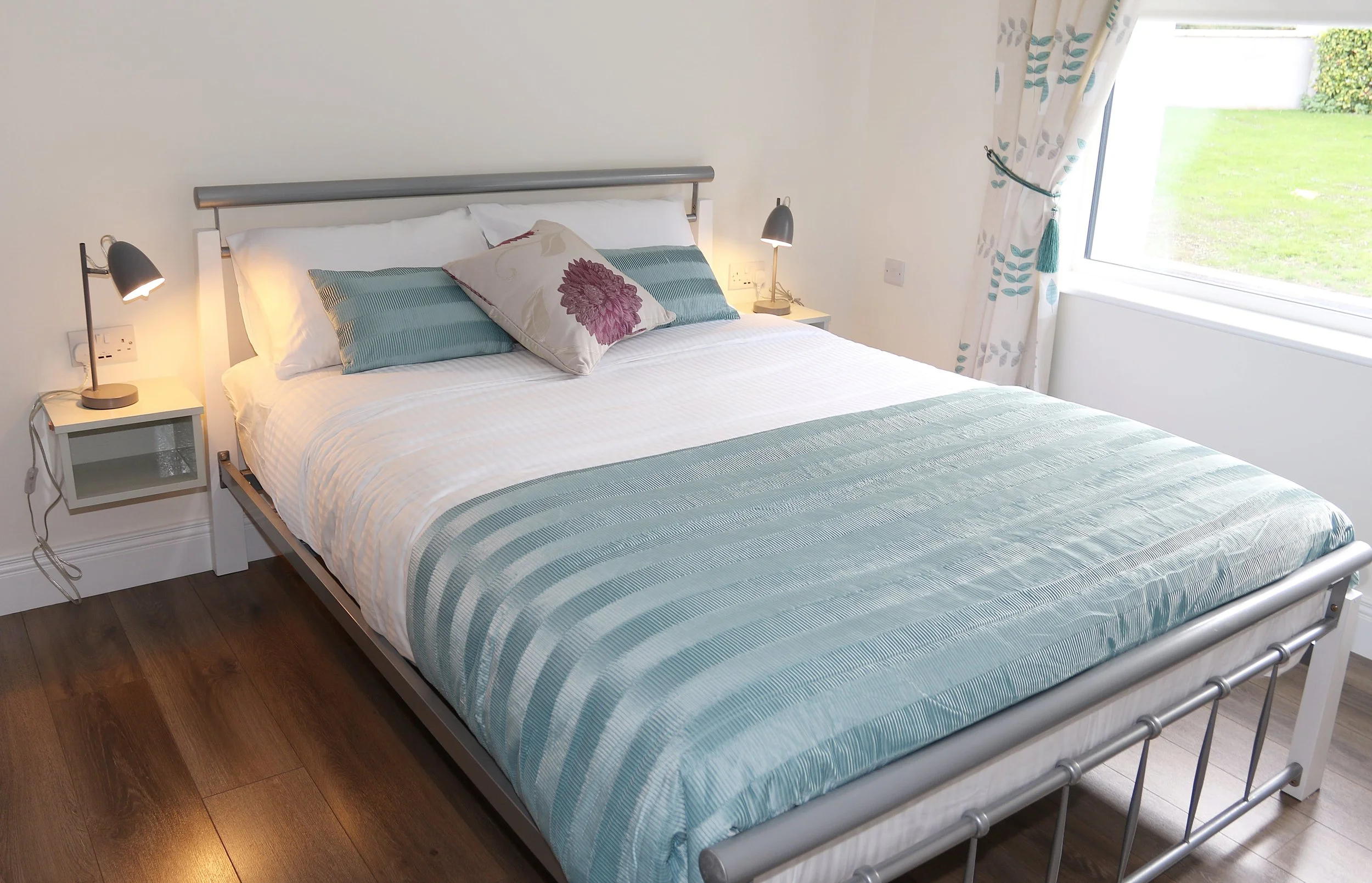 A neatly made bed with a striped duvet and pillows, including one with a floral design, in a room with a window and curtains showing a grassy outdoor view. There are two bedside tables with lamps.