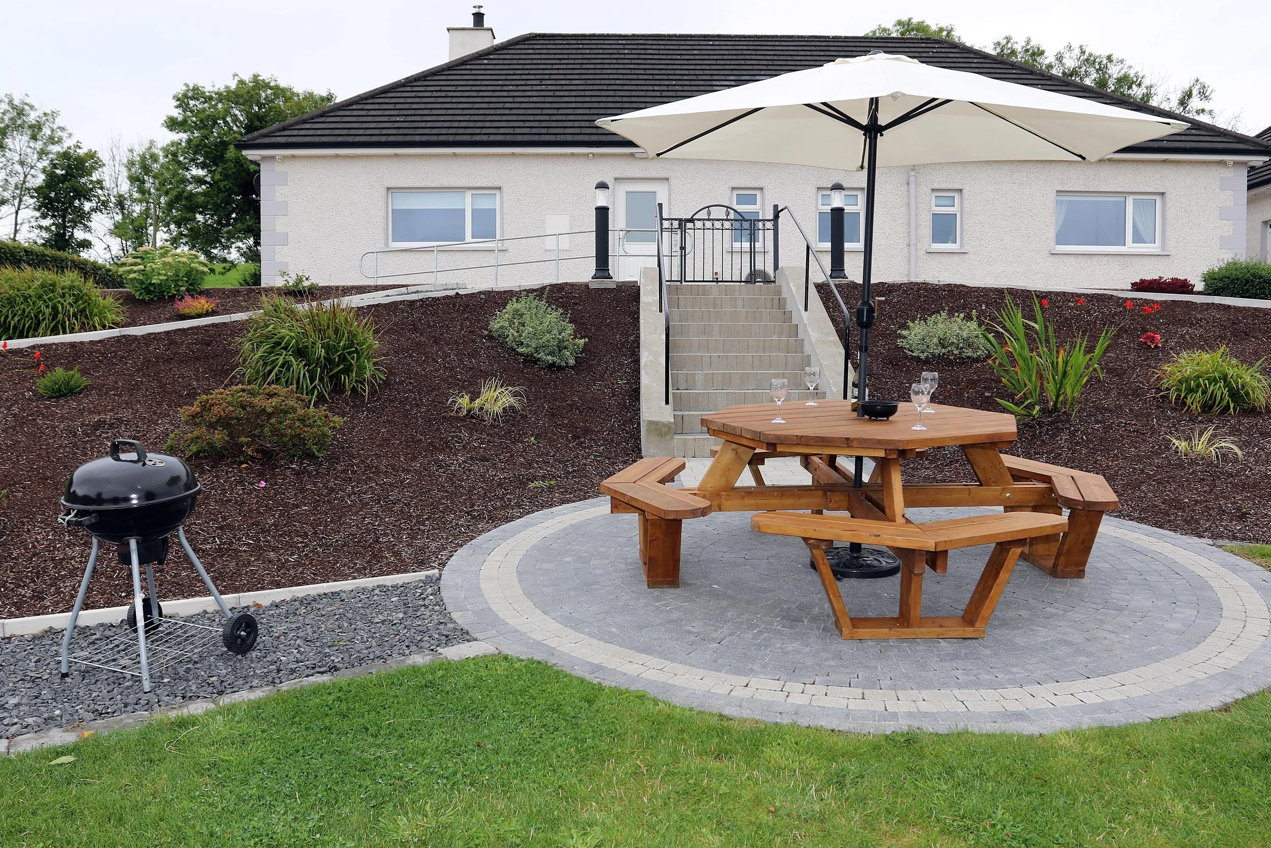 Backyard patio with umbrella, picnic table, grill, and landscaped flower beds in front of a house with white walls and gray roof.