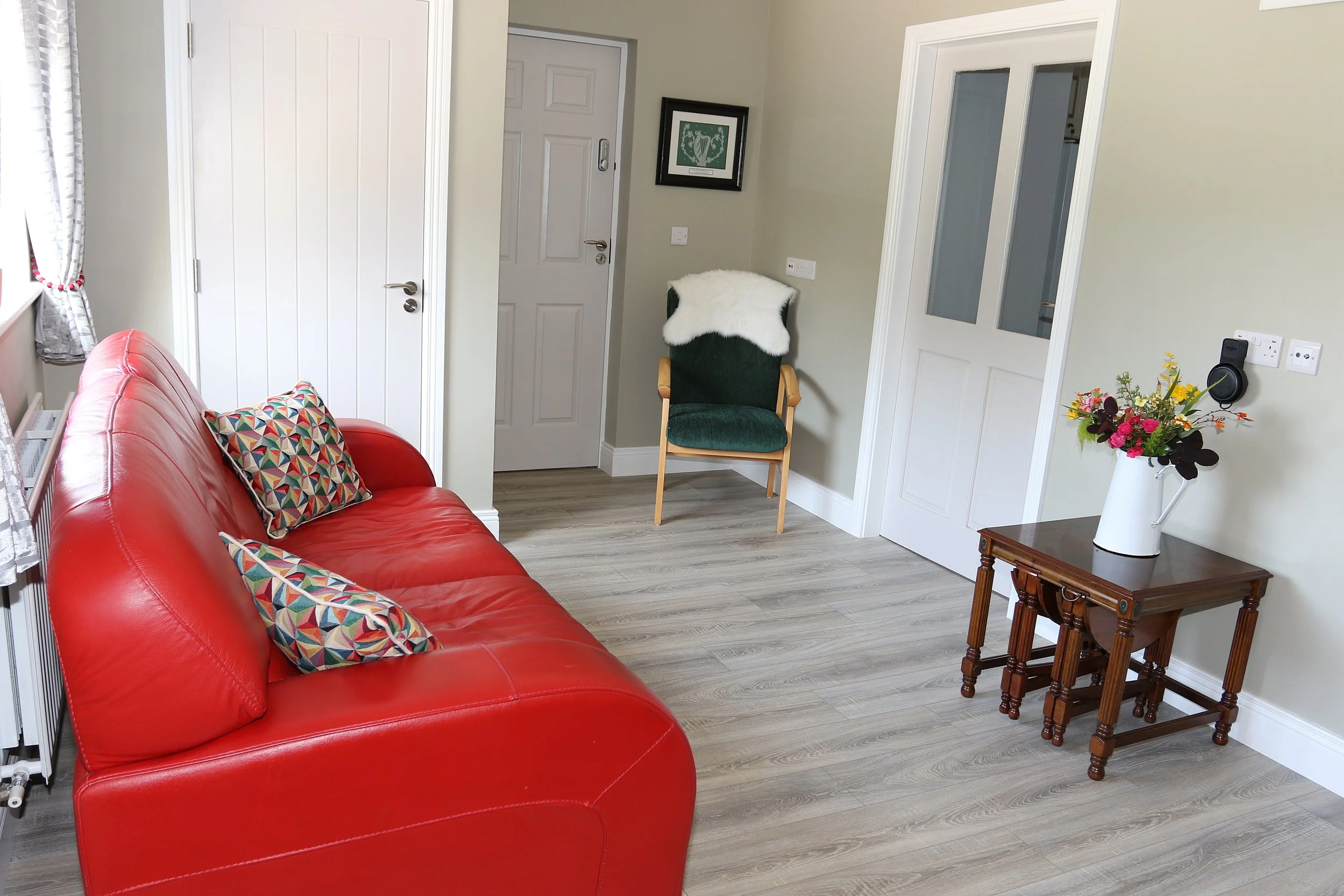 Interior of a living room with a red leather sofa, a patterned pillow, a green armchair with a white fuzzy throw, a small wooden table with a vase of colorful flowers, and a framed picture on the wall near a white door.