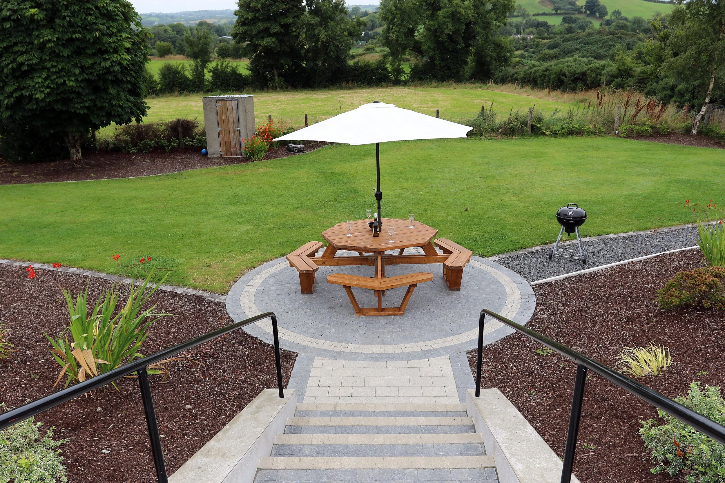 View from top of stairs looking down at a backyard with a paved circular patio, picnic table with an umbrella, a small grill, and a landscaped garden. Beyond is a grassy yard and trees with hills in the distance.