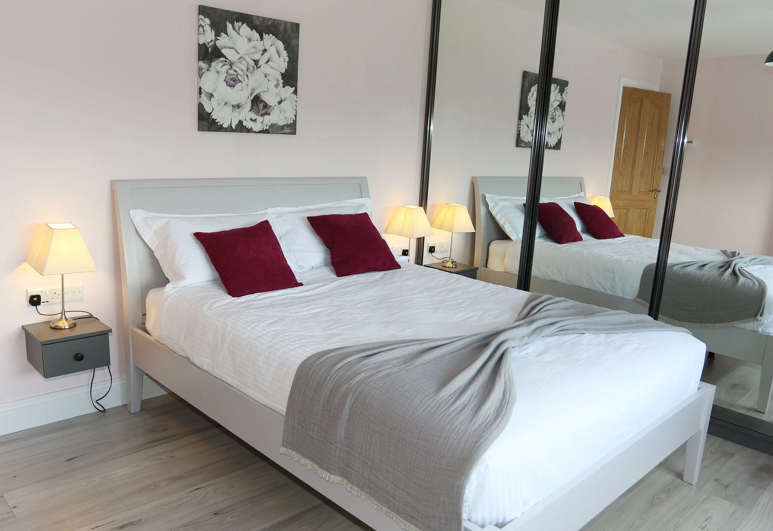 A neatly made bed with white bedding, two burgundy pillows, and a gray throw blanket. Two bedside lamps are on small tables, and a large mirror reflects part of the room. A floral painting hangs above the bed. The room has light-colored walls and laminate flooring.