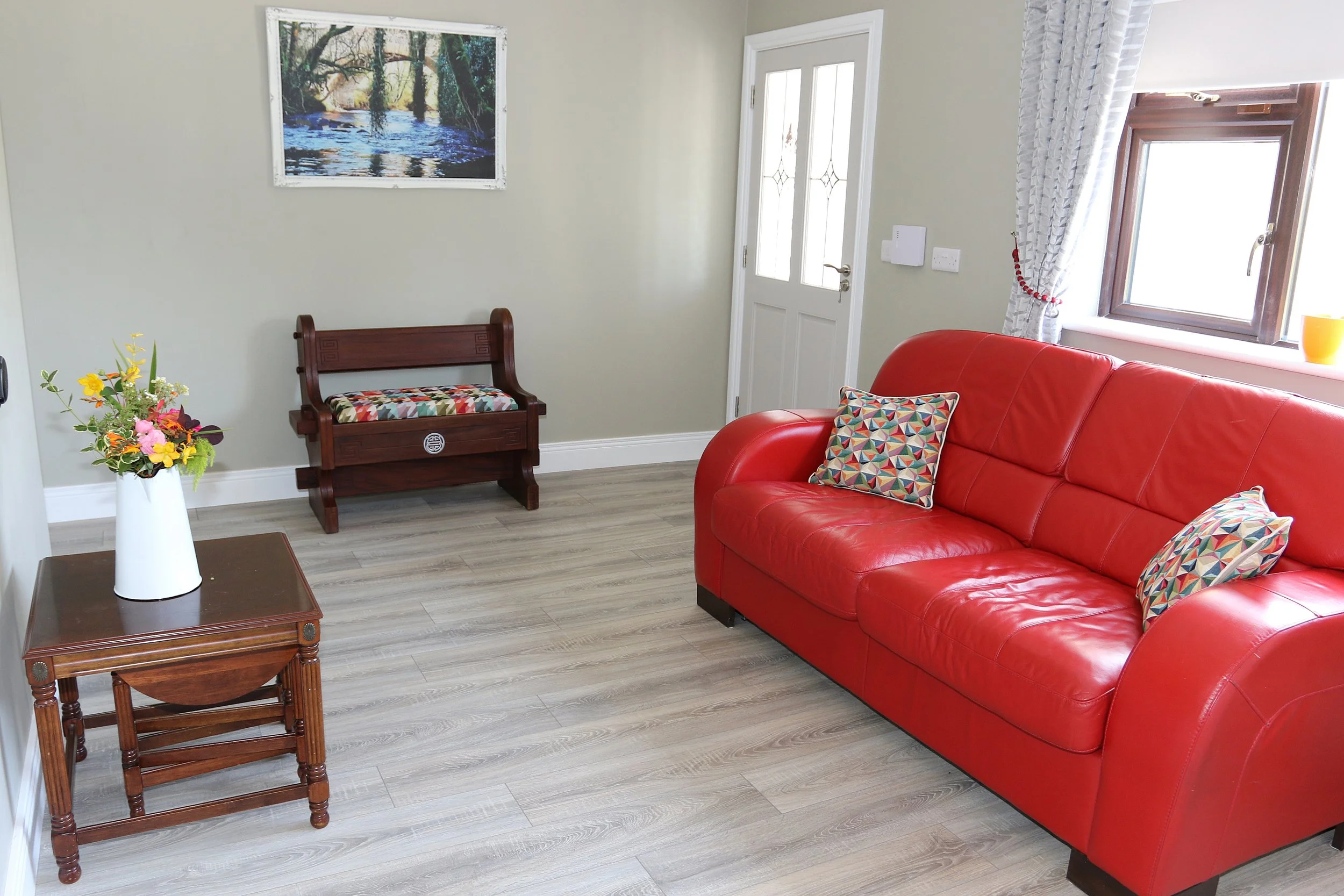 Living room with a red leather sofa, patterned pillows, wooden coffee table, small brown stool, white vase with flowers, framed river scene picture on the wall, white door, and window with white curtains.