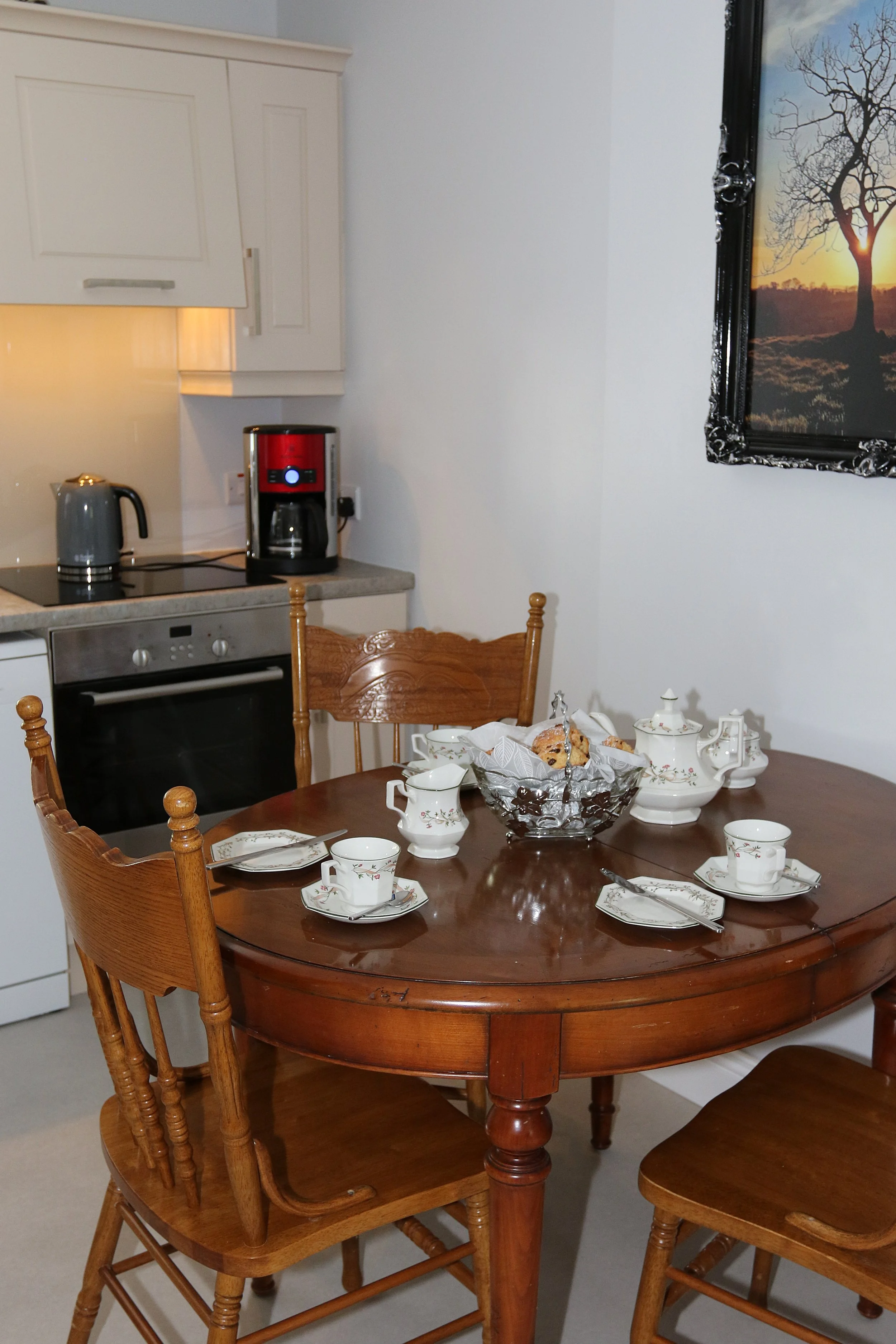 A wooden dining table set with fine china, teapots, and cups, surrounded by wooden chairs, in a kitchen with white cabinets and a wall-mounted framed picture of a tree at sunset.