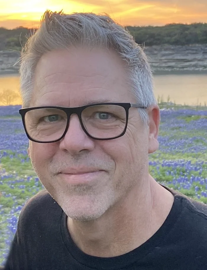 A man with glasses smiling in a field of purple flowers near a body of water during sunset.