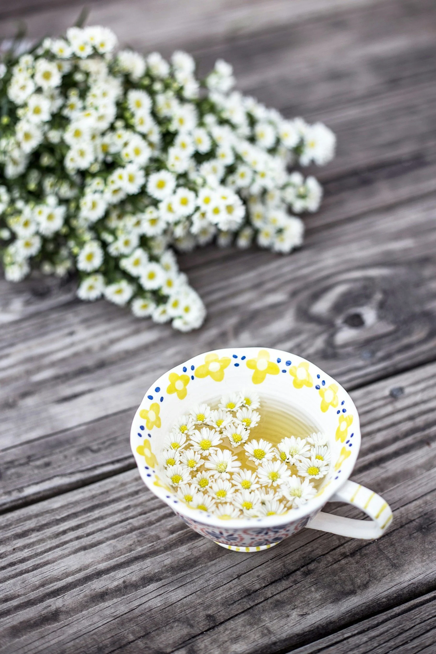 A ceramic mug with a floral pattern filled with chamomile tea and floating chamomile flowers, on a weathered wooden surface, with a blurred bouquet of white daisies in the background.