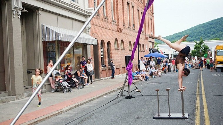 a circus performer balances on their hands in the foreground - one leg bent. they are performing on a downtown street with a mountain in the background.