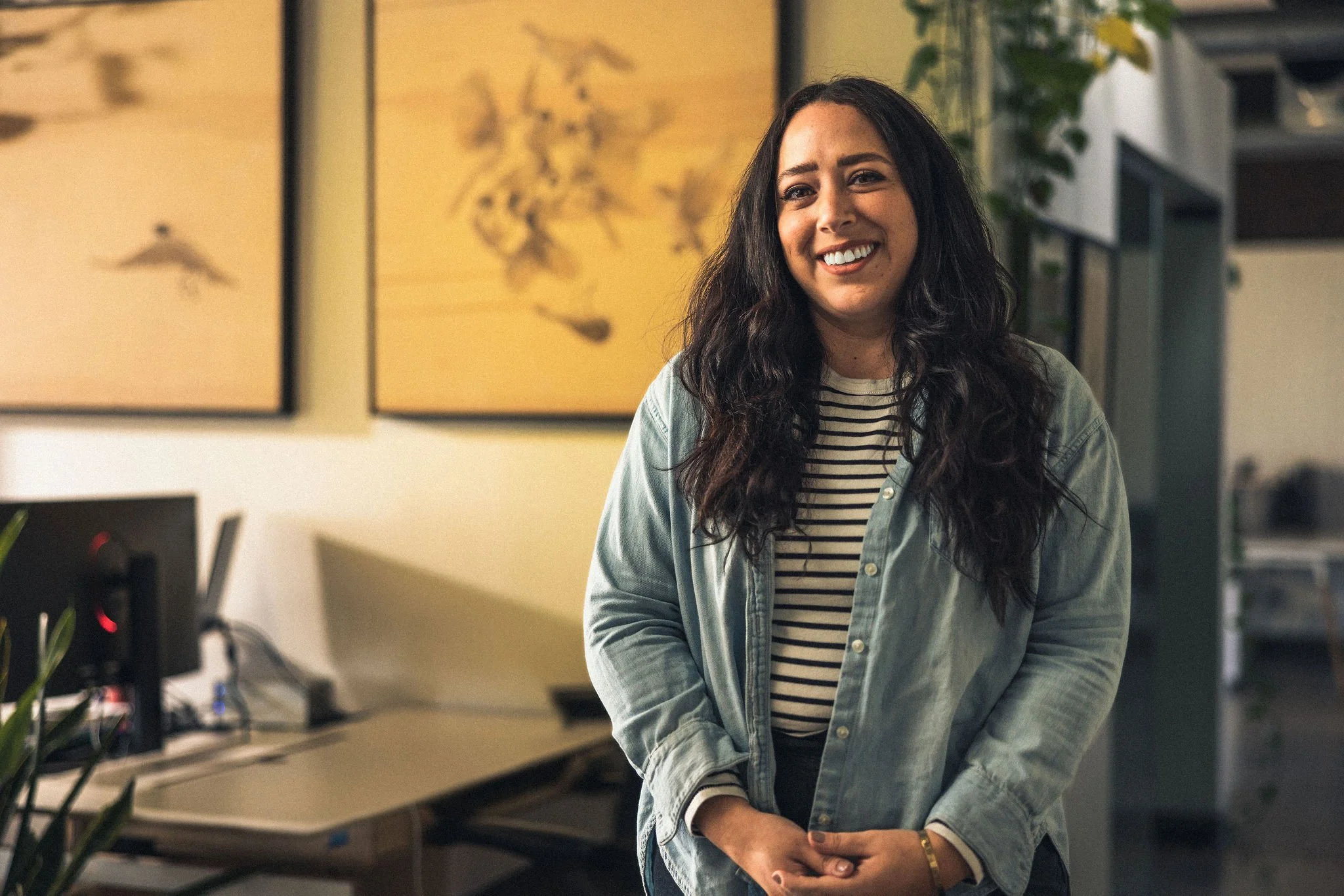 A woman with long dark hair, smiling, standing in an office environment with a yellow painting and green plant in the background.