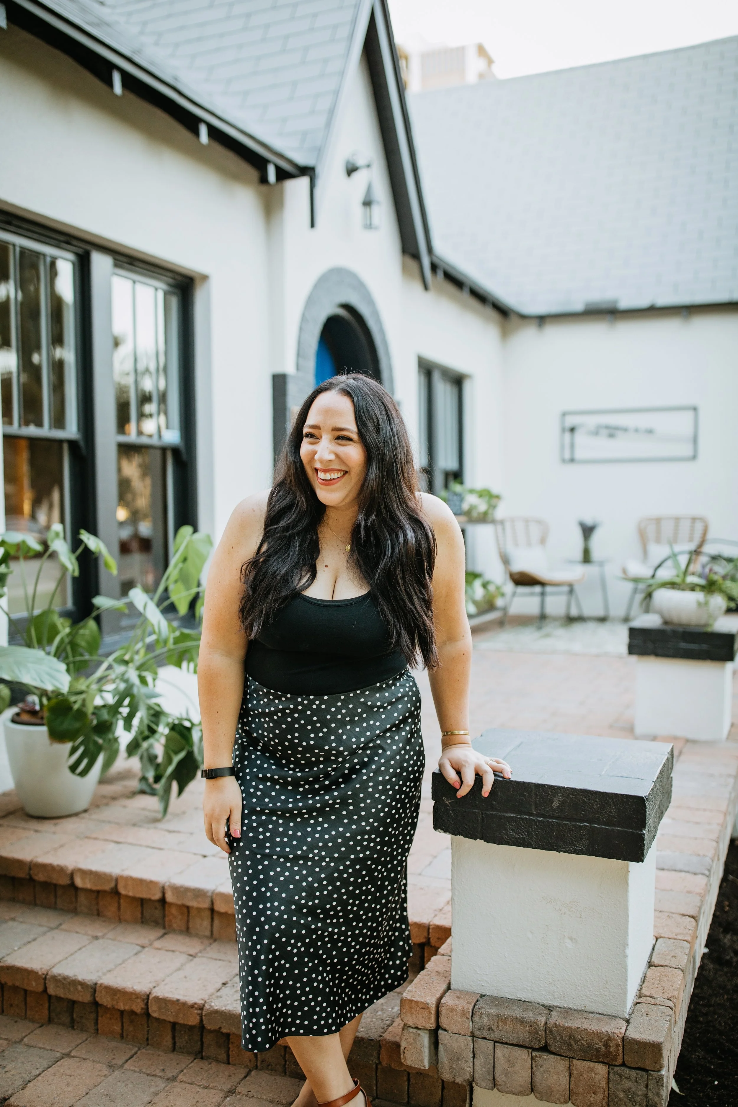 A woman with long dark hair smiling outdoors, standing on brick steps next to a white pillar in a residential backyard or patio area with plants and house in the background.
