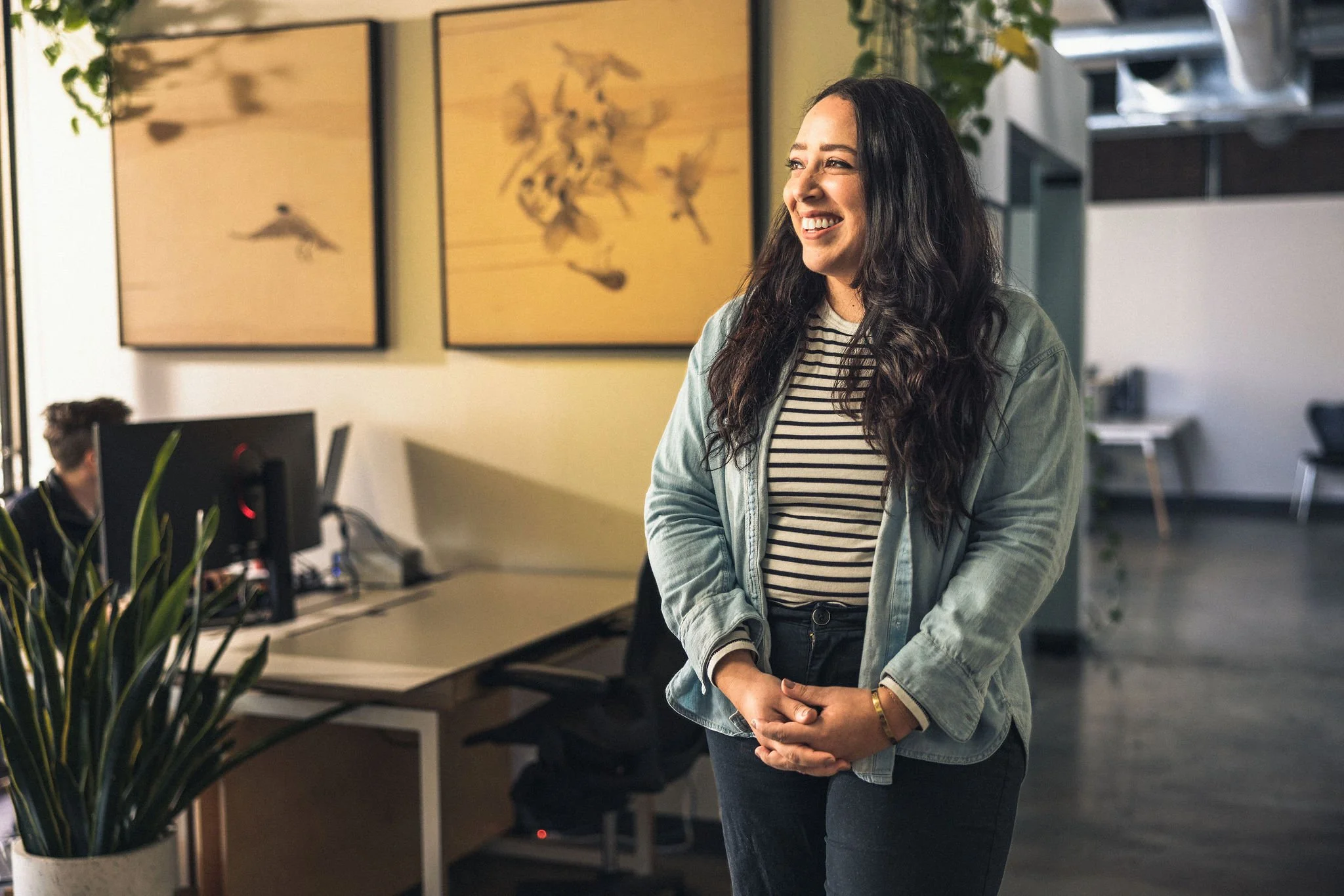 A woman with long dark hair smiling, standing in an office environment with computer monitors and artwork on the wall.
