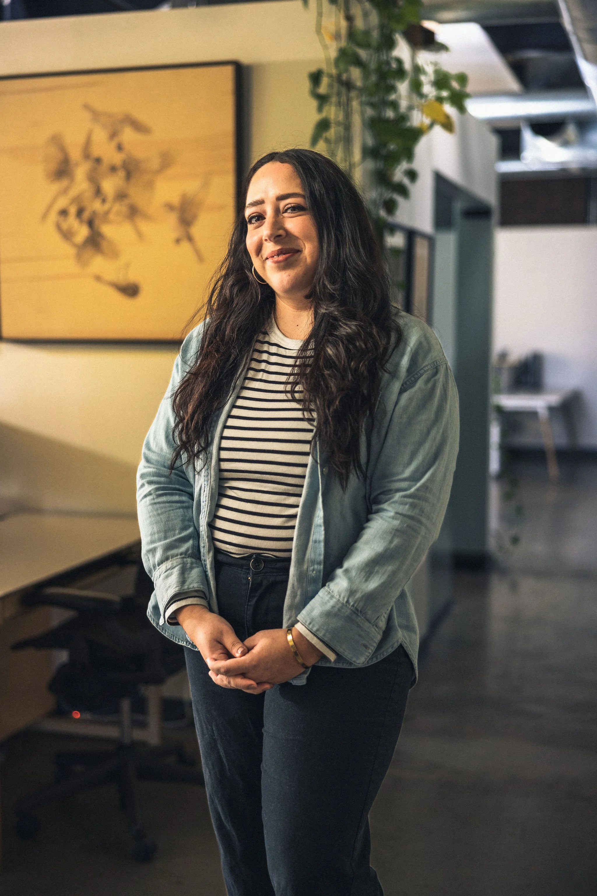 A woman with long dark hair, wearing a striped shirt and a denim jacket, standing in an office environment with a blurred background of a desk and a plant.
