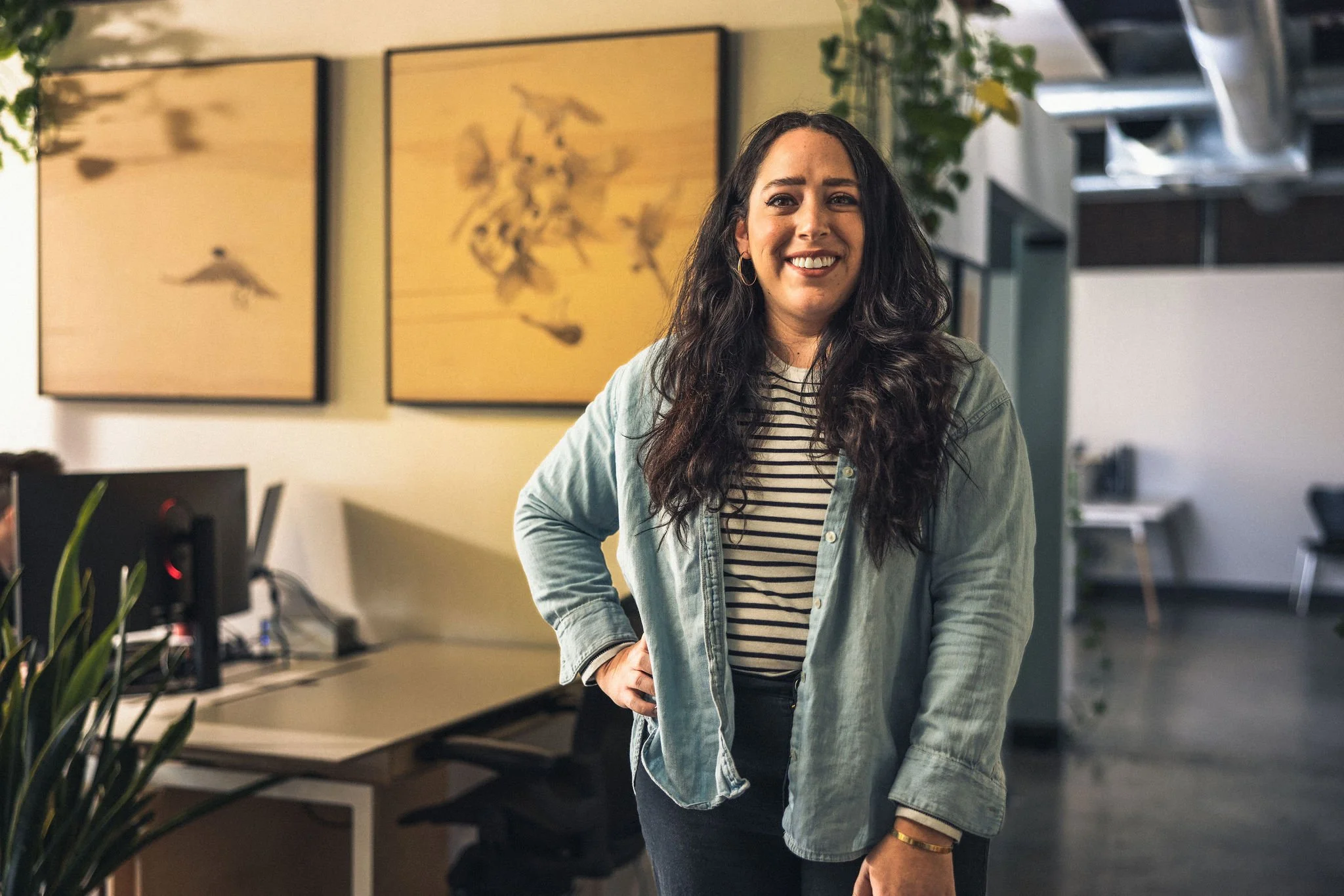 Woman with long dark hair, wearing a striped shirt and denim jacket, smiling in an office setting with art on the walls and plants around.