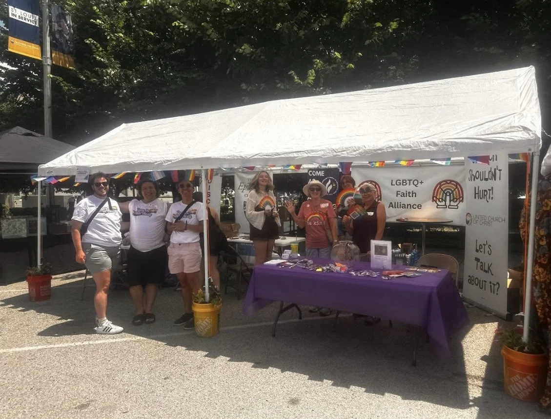 LGBTQ+ Faith Alliance members smiling under a tent with a table full of community resources.