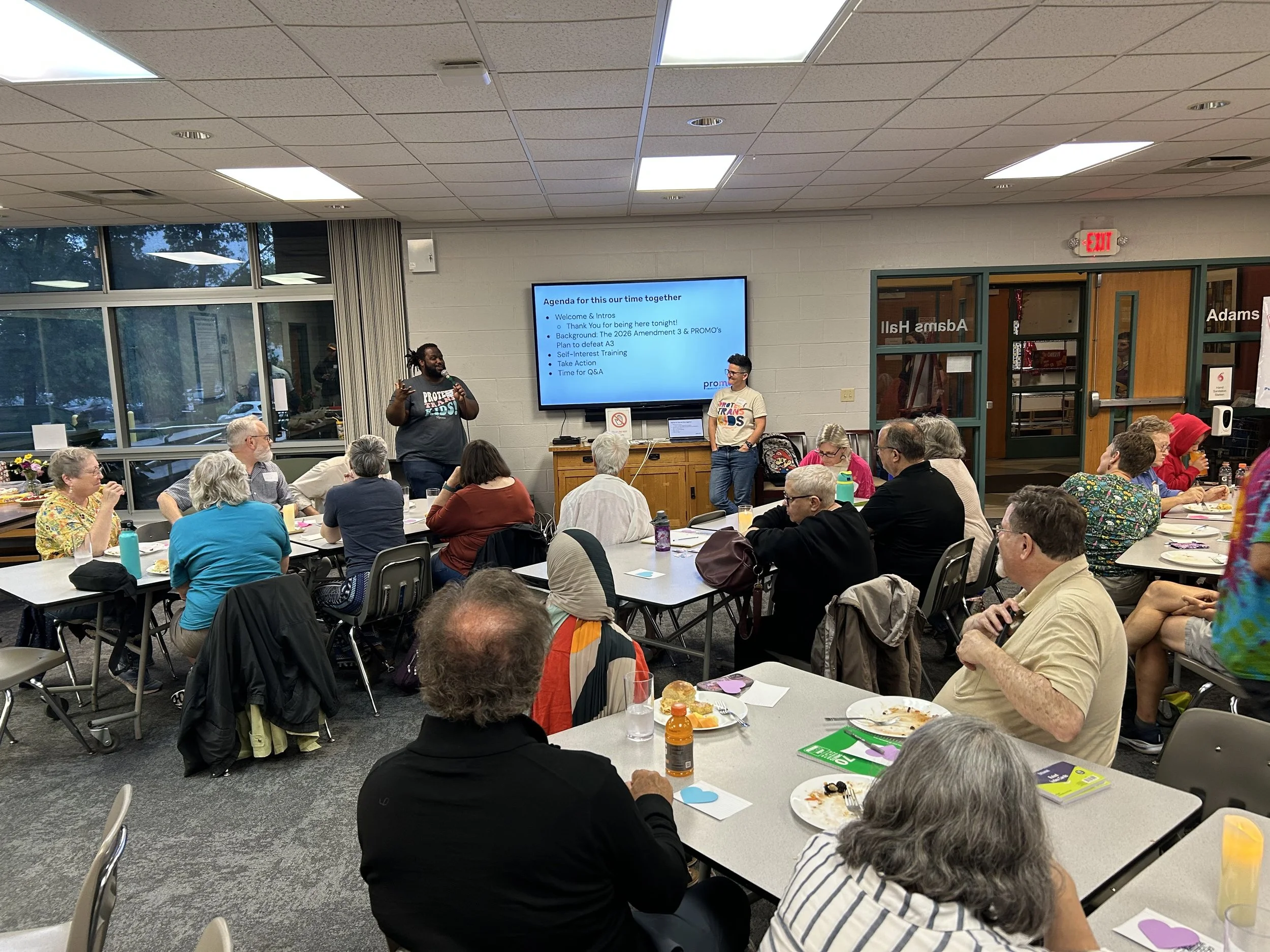 Two people standing at the front of a room, presenting to a group of people sitting at tables.