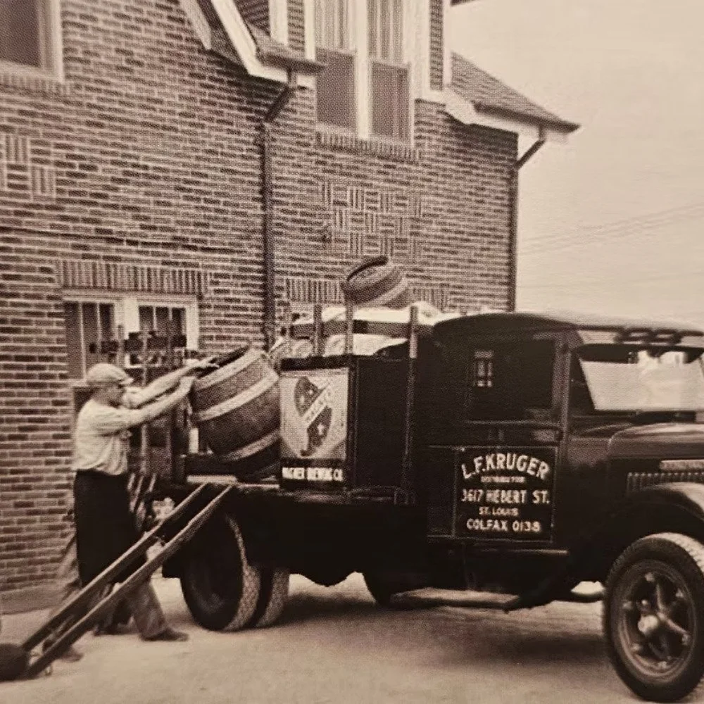 A man load a vintage pickup truck with barrels and a sign says 'L. F. Kruger'.