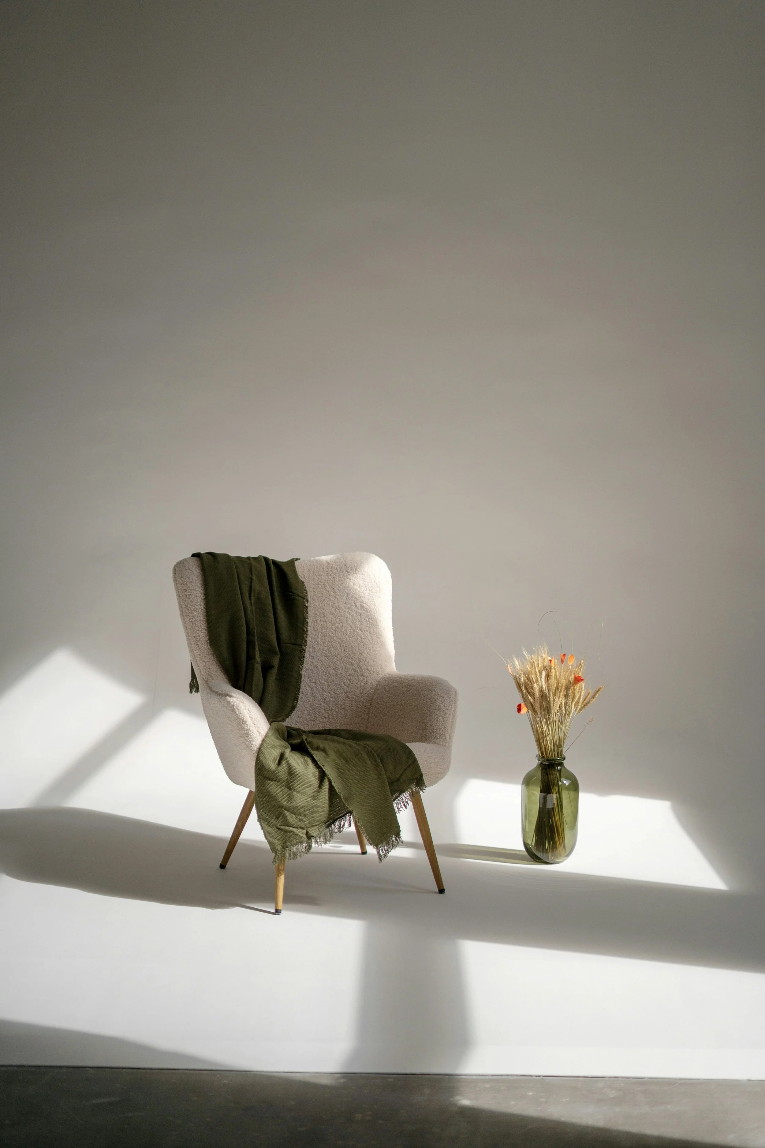 A beige upholstered armchair with wooden legs, draped with a green blanket. Next to it, a tall glass vase with dried orange and beige flowers, set against a plain light grey wall with lighting casting shadows.