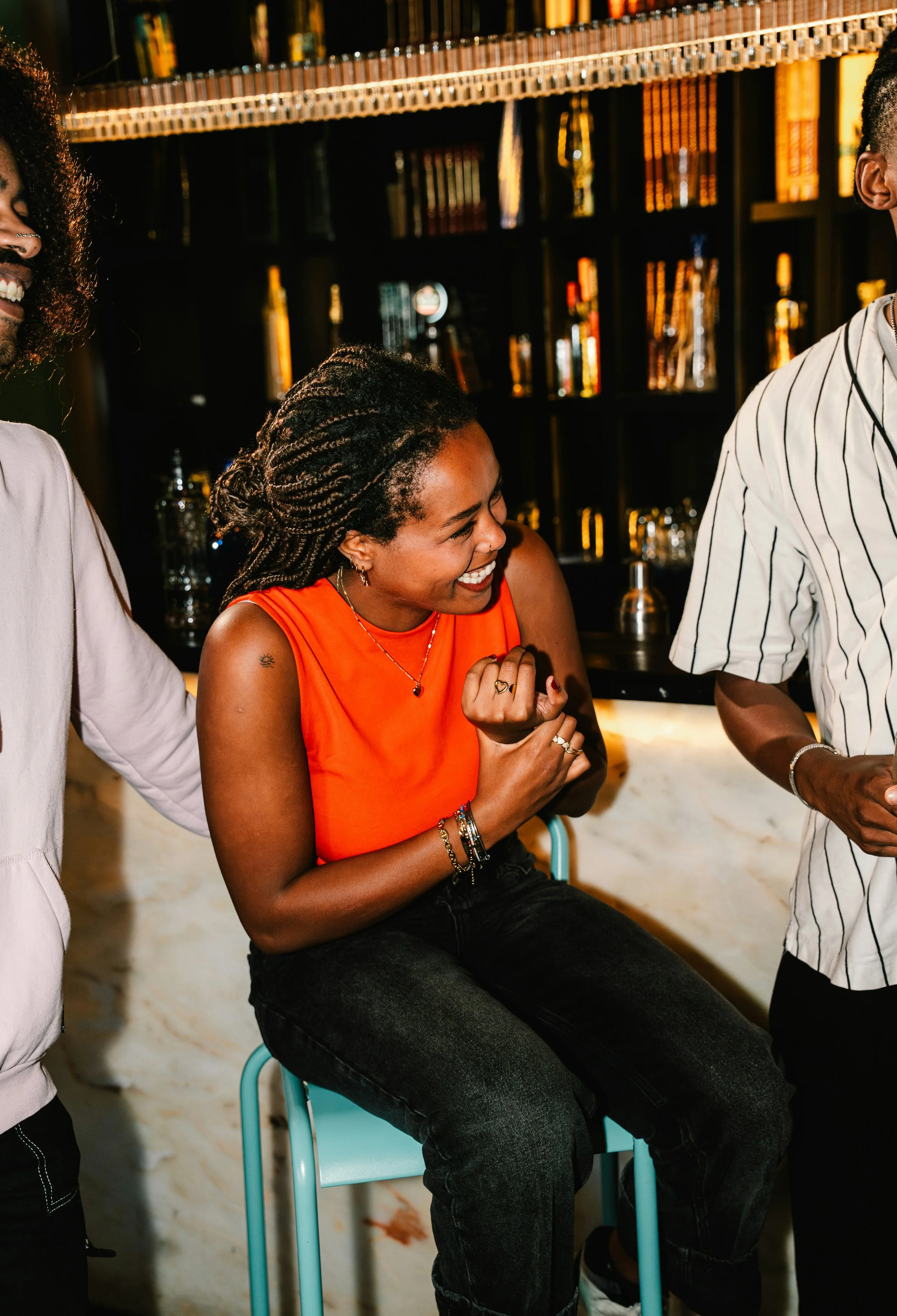 A woman with braided hair wearing an orange sleeveless top, sitting on a light blue chair, laughing and smiling while chatting with a group of people in a bar or lounge setting with a black bookshelf and warm lighting in the background.