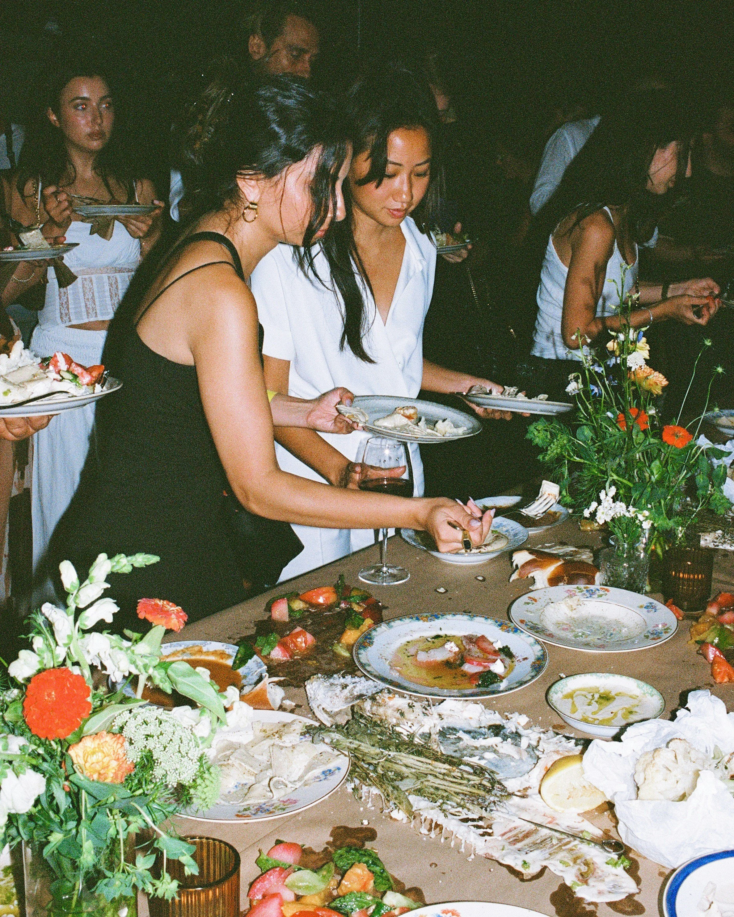 A group of women and a man selecting desserts from a buffet table filled with various cakes, fruits, and decorations at a party or celebration.