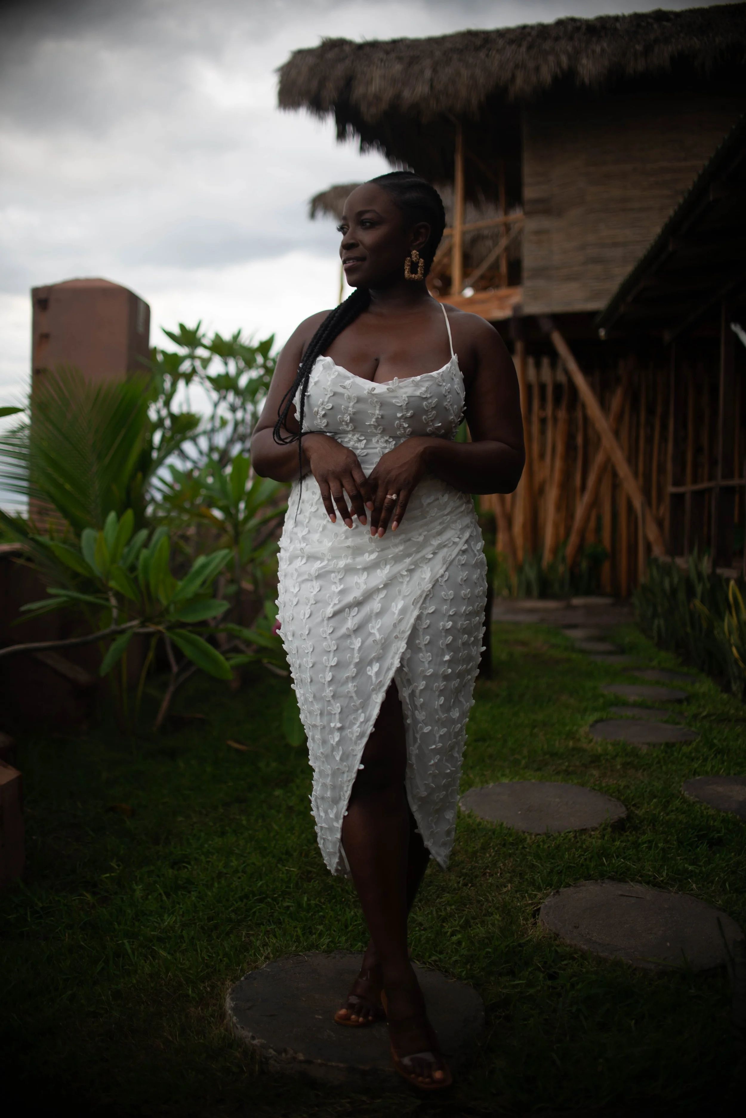 A woman in a white embroidered dress with spaghetti straps and large gold earrings stands outdoors near a traditional bamboo house with a thatched roof. She has braided hair and is standing on a stone pathway surrounded by green plants, with cloudy skies overhead.