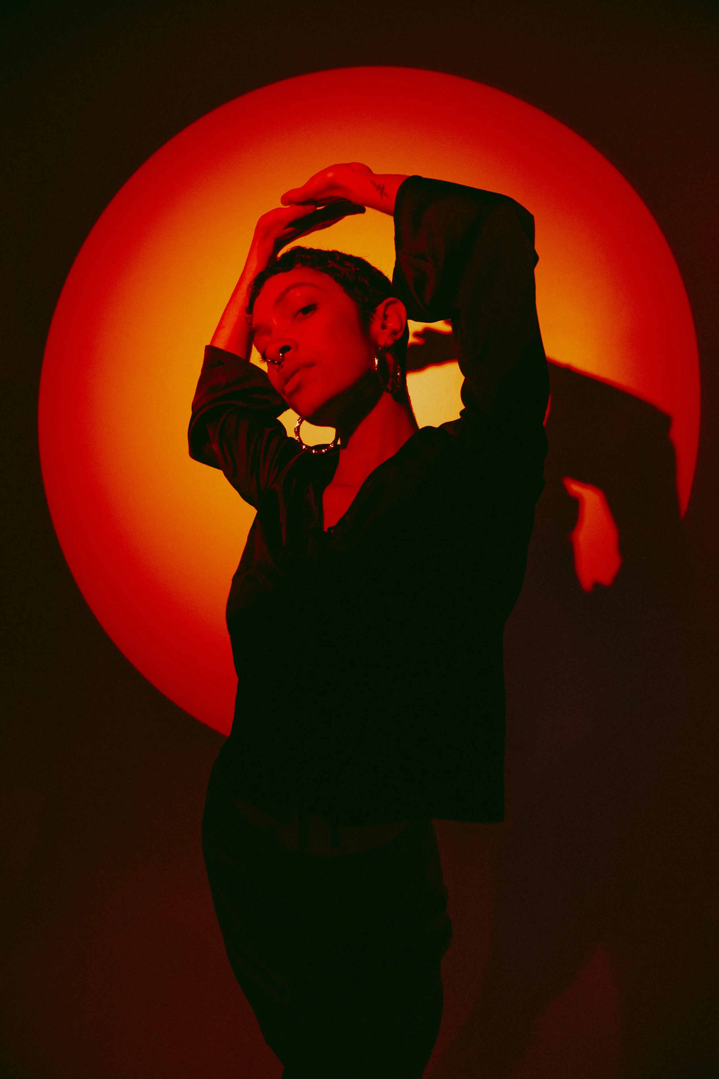 A person with short dark hair, wearing dark clothing, posing with their arms raised above their head, against a bright red and orange circular background, creating a striking silhouette.