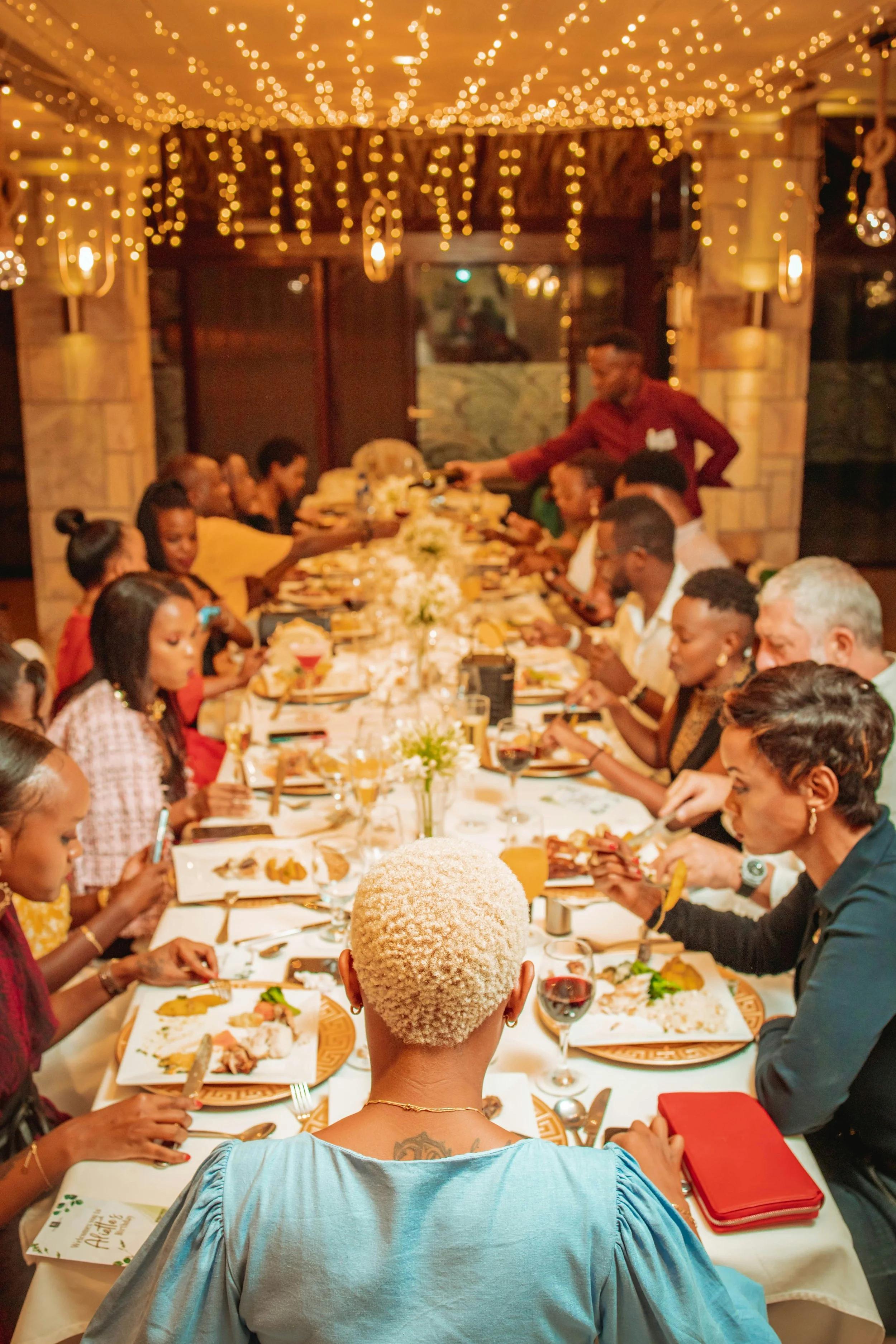A group of diverse people gathered around a long dinner table, enjoying a meal, with a woman with short, curly white hair sitting at the head of the table in the foreground. The room is decorated with warm lighting and string lights hanging from the ceiling.