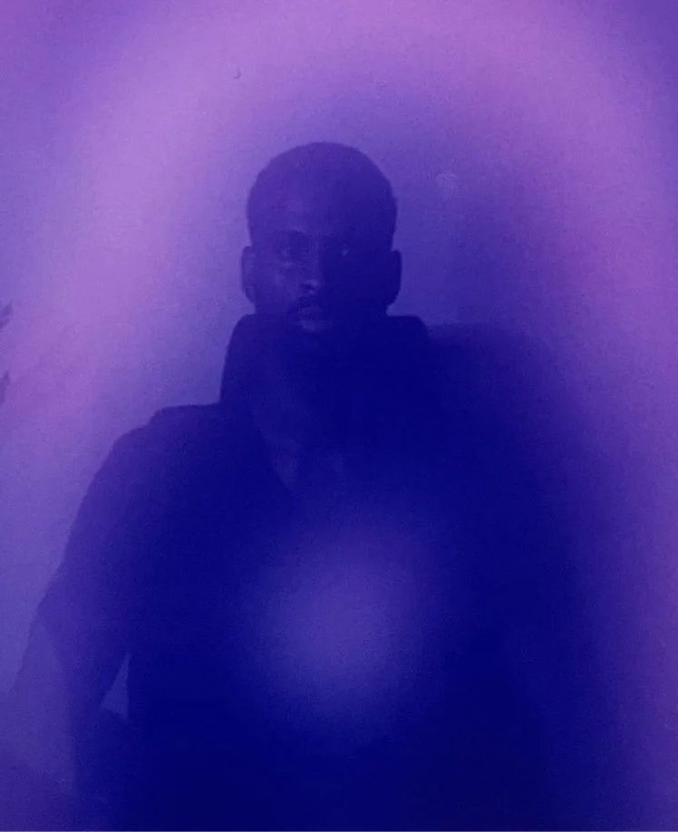 A person taking a mirror selfie in a dimly lit room with purple lighting, wearing glasses and a dark hoodie.