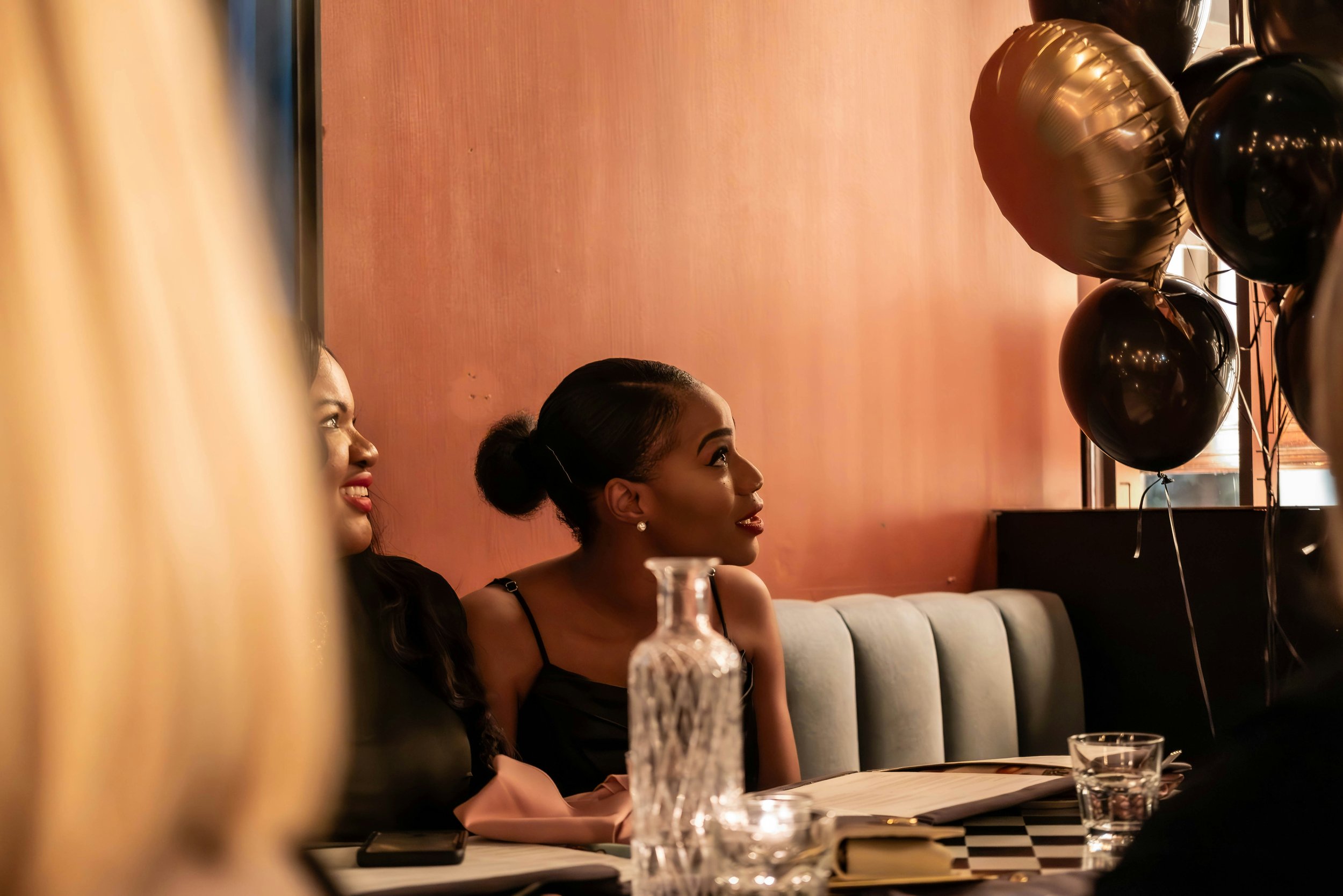 Two women sit at a decorated table with balloons in the background, enjoying a celebration or party.