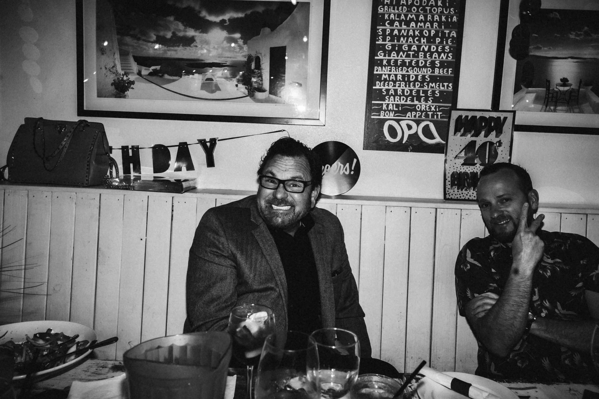 Two men sitting at a dining table in a restaurant. The man on the left has glasses and a beard, smiling at the camera, wearing a blazer. The man on the right has a goatee, smiling, with his hand near his face, wearing a patterned shirt. The table has glasses with drinks, a bowl of food, and utensils. The background features framed artwork and a menu board on the wall.