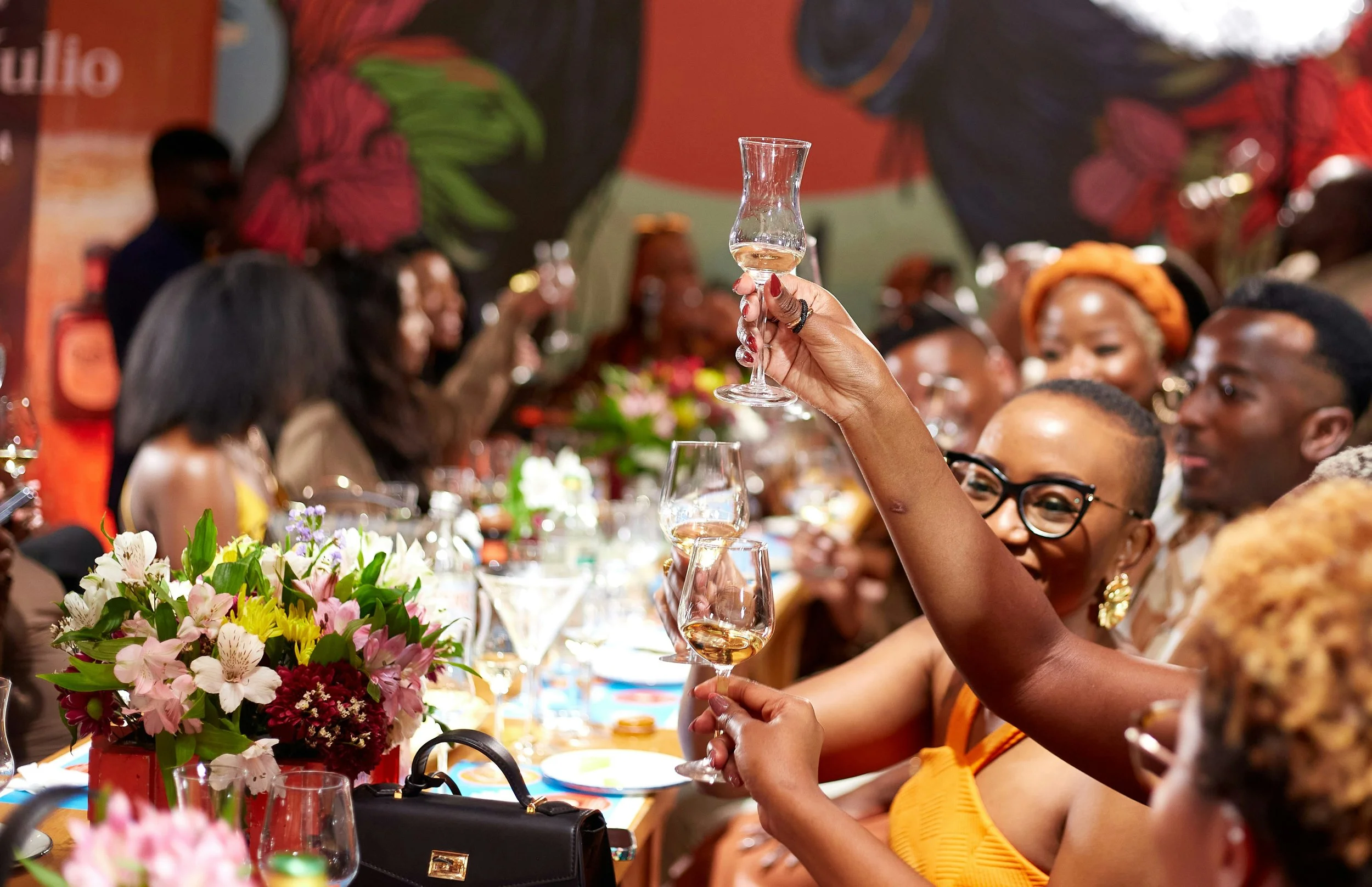 A group of people at a celebration or party, raising glasses of wine for a toast, with floral centerpieces on the table and colorful decorations in the background.