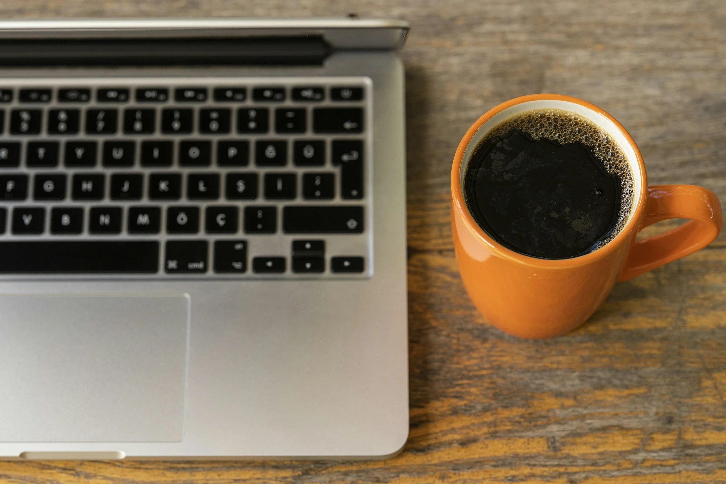 Top view of a silver laptop and an orange mug filled with black coffee on a wooden table.