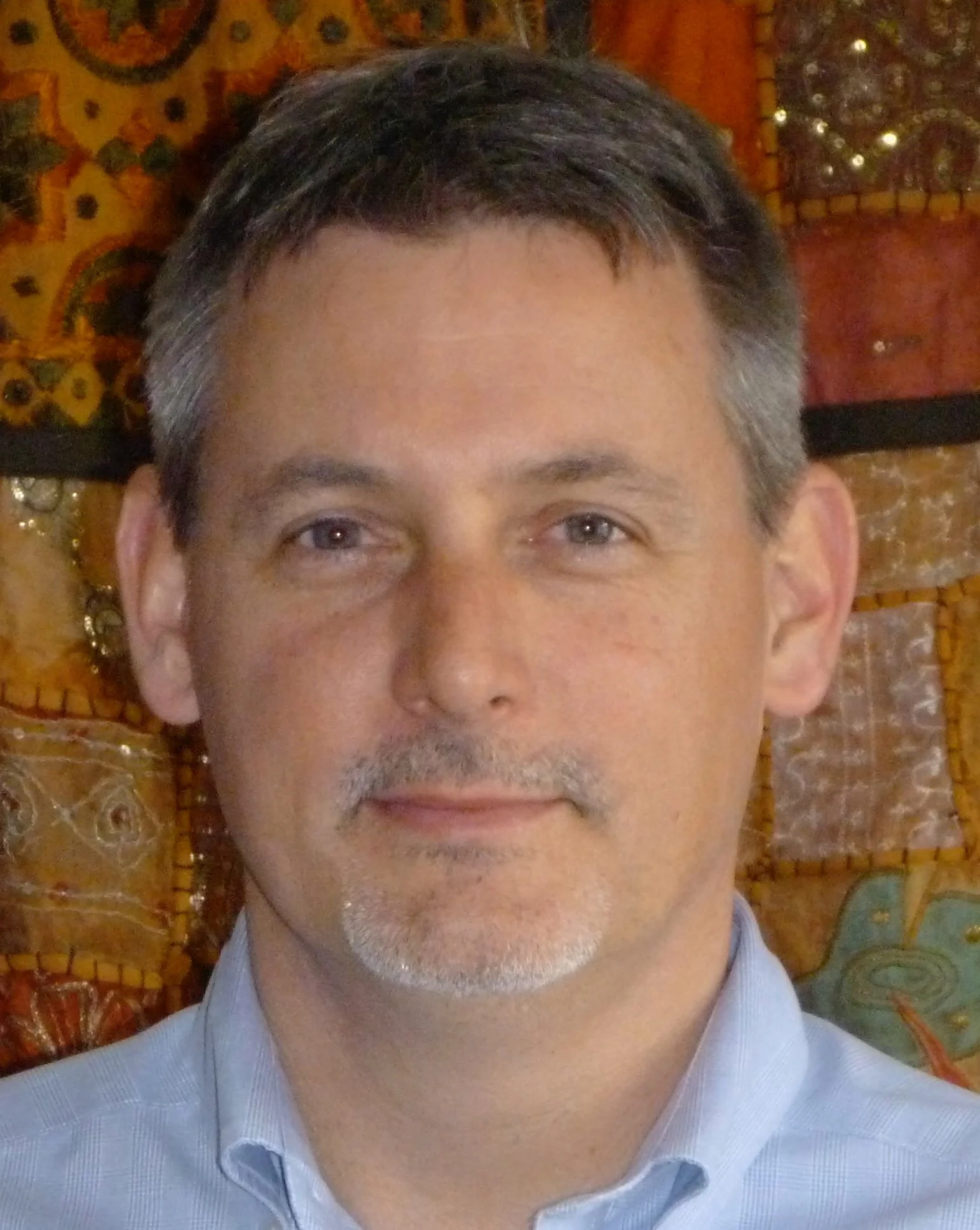 Close-up of Scot Cooper RP with graying hair and a goatee, wearing a light blue collared shirt, in front of a decorative background with orange and gold patterns.