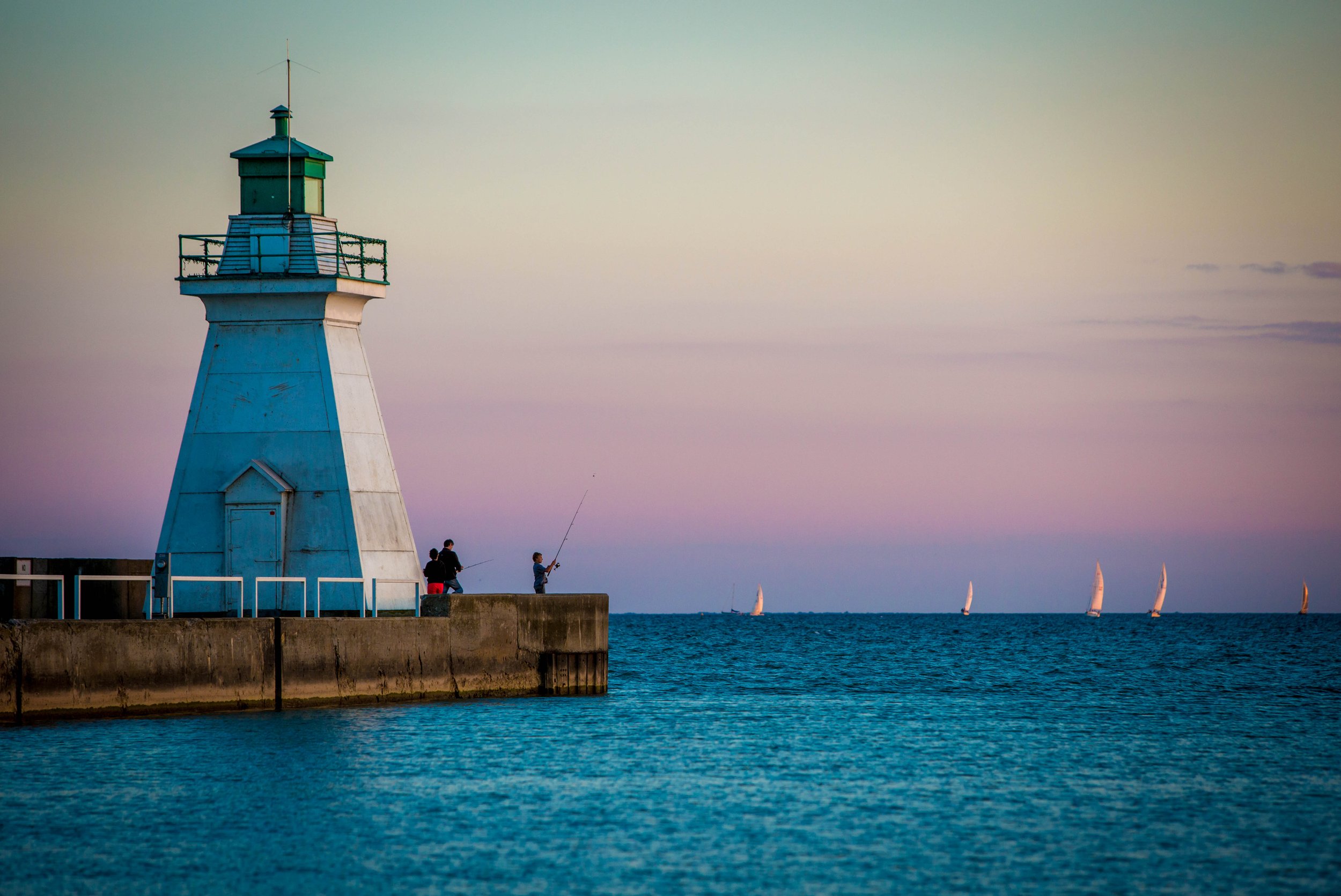 A lighthouse on a pier with several children fishing, sailboats in the water, and a clear sky at sunset.