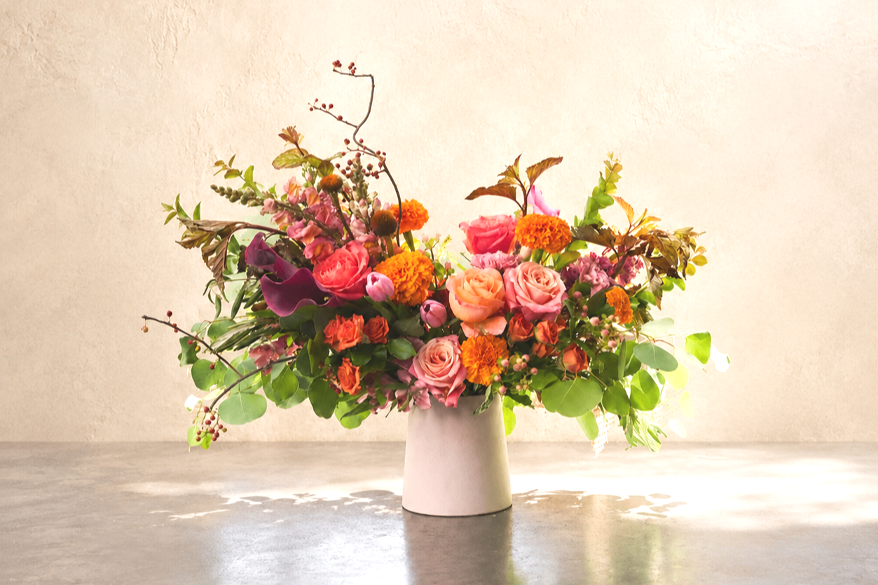A colorful bouquet of flowers in a white vase, featuring pink roses, orange marigolds, dark purple calla lilies, and various greenery on a gray surface with a light beige background.