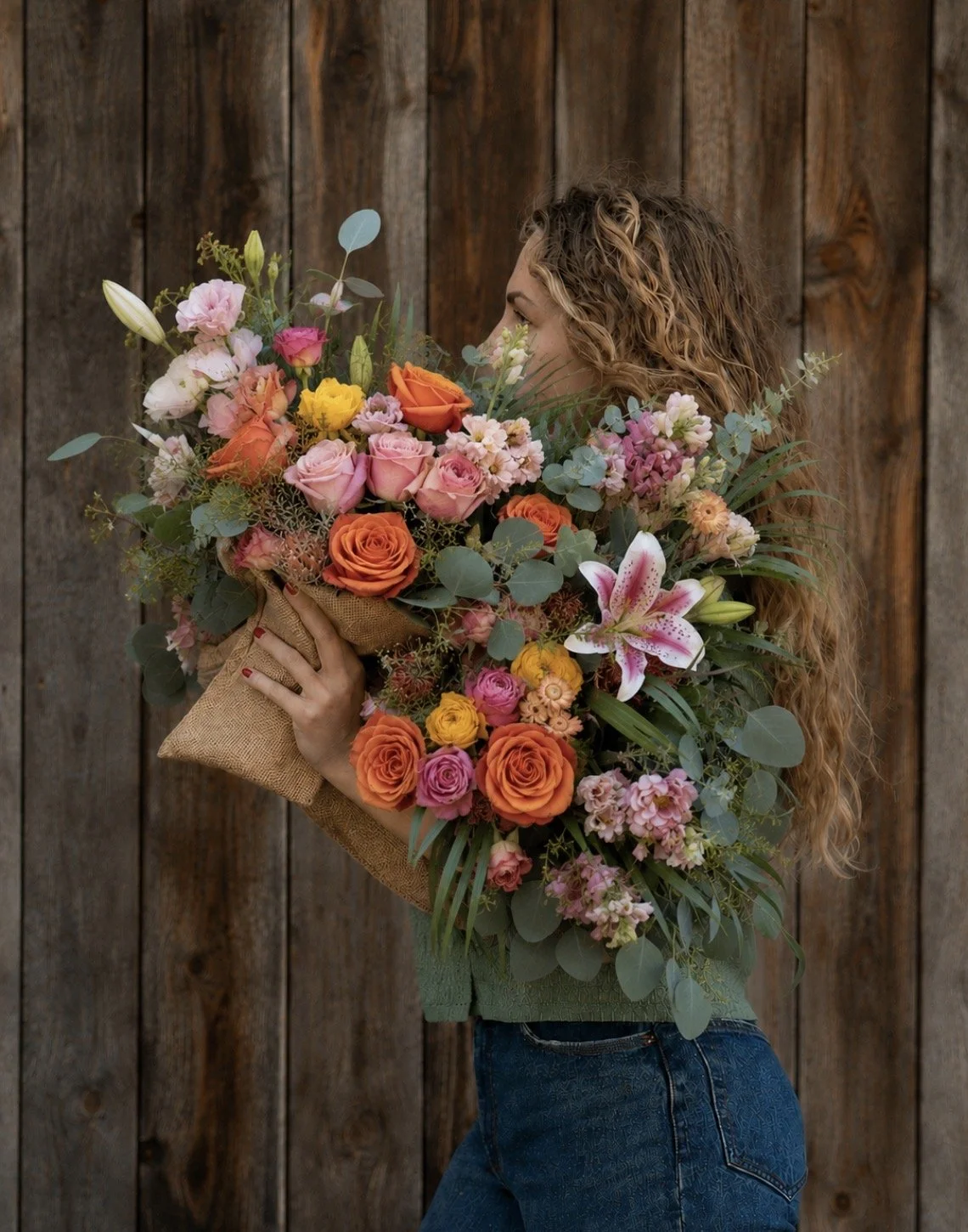 A woman with curly hair holding a large bouquet of colorful flowers in front of a wooden wall.