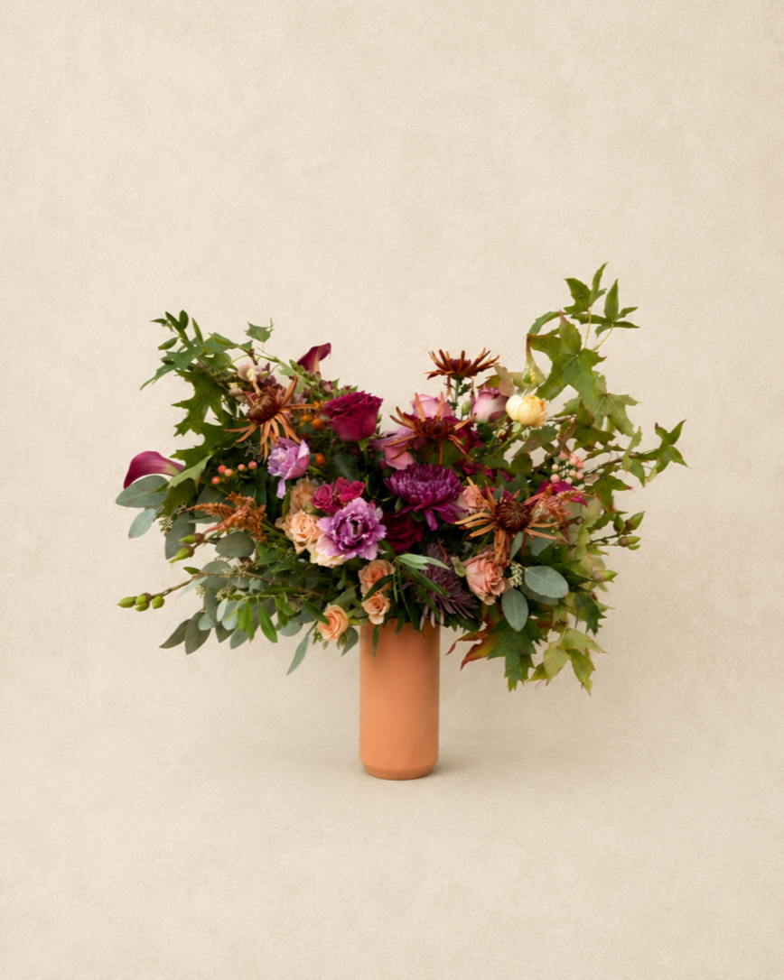 A bouquet of mixed flowers in a tan ceramic vase against a neutral background.