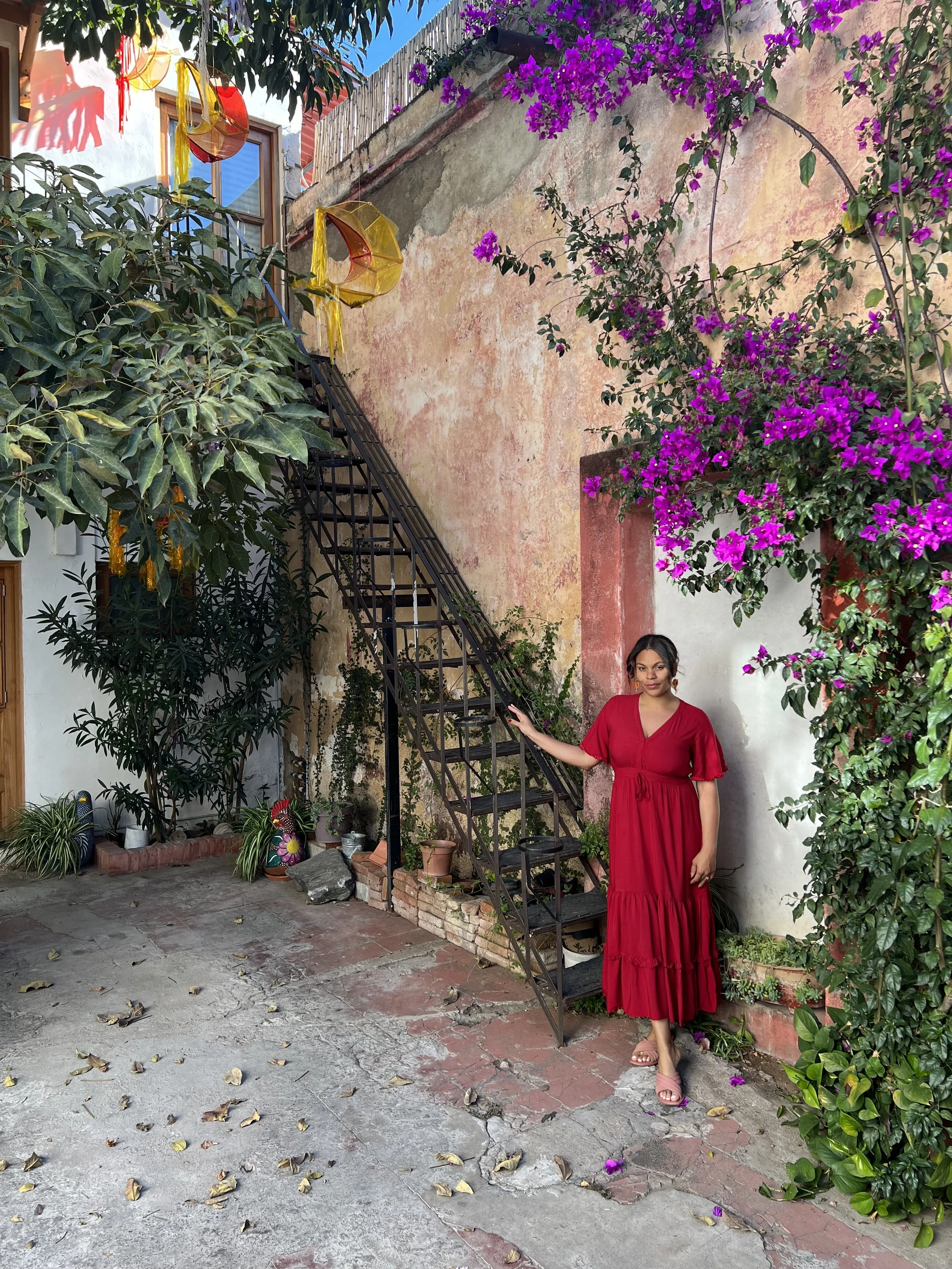 A woman in a red dress standing in a courtyard with colorful flowers and plants, holding a metal staircase railing, with a weathered stucco wall and hanging decorations in the background.