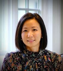 Portrait of a woman with black hair, wearing a floral-patterned blouse, standing in front of a window with white curtains.