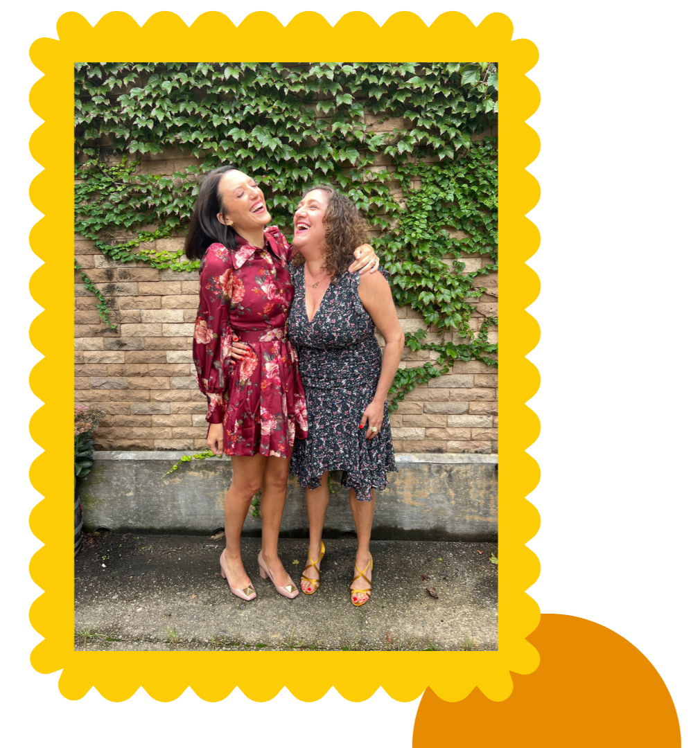 Two women standing outside in front of a brick wall covered in ivy, laughing and embracing each other.