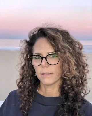 Woman with curly hair and glasses outdoors at sunset or sunrise.