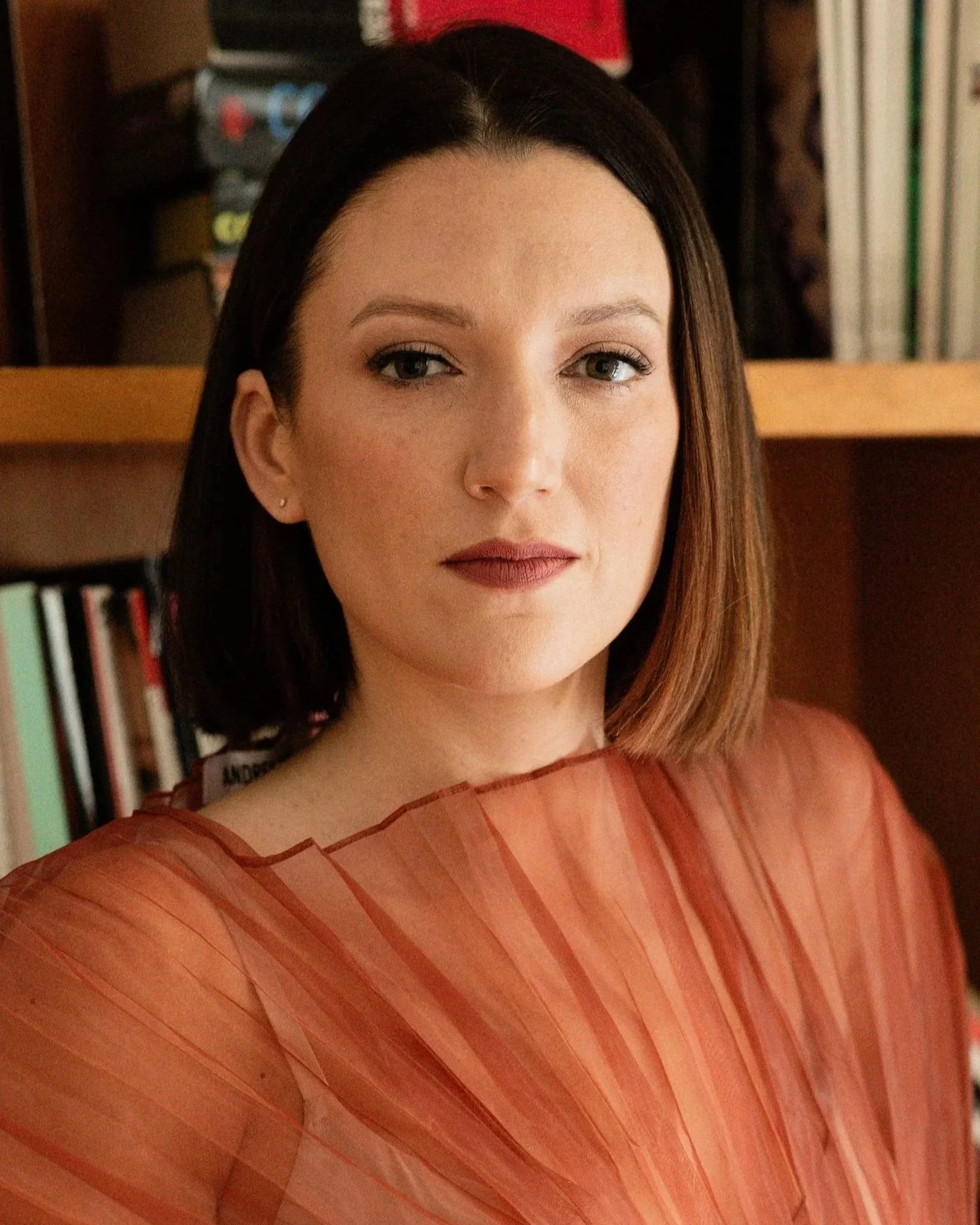 Close-up of a woman's face with black hair, wearing makeup including lipstick, and a sheer, orange, striped fabric covering her shoulder, with books and boxes in the background.